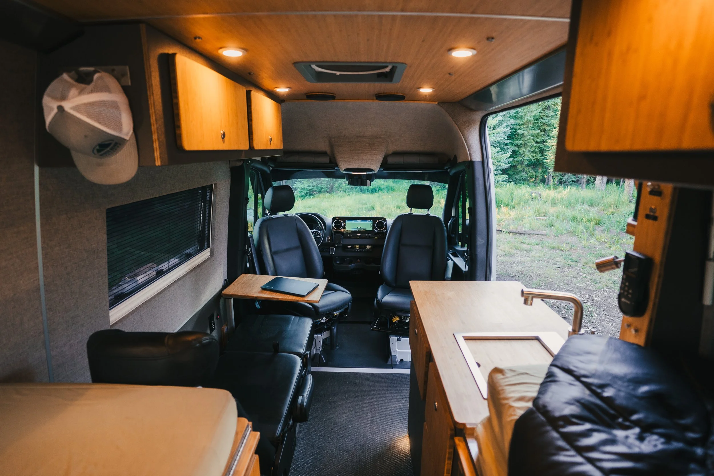 Interior of a camper van with two front seats, wooden cabinets, a small table, window, and a kitchen counter with a sink, overlooking a green outdoor landscape.