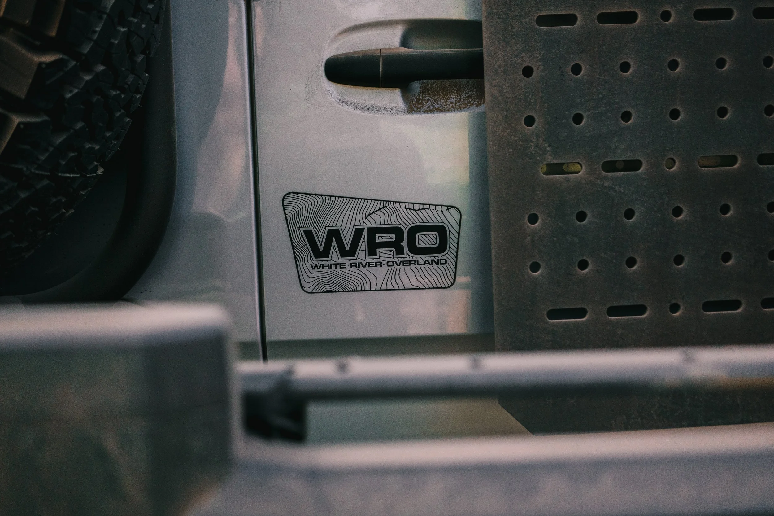 Close-up of a sticker reading 'WR 0 WHITE RIVER OVERLAND' on a vehicle, with part of a tire, a handlebar, and metal structure visible.