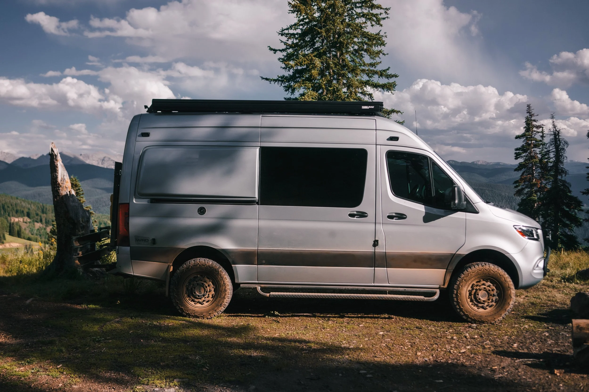 A silver camper van parked in a mountainous landscape with green trees, mountains in the background, and partly cloudy skies.