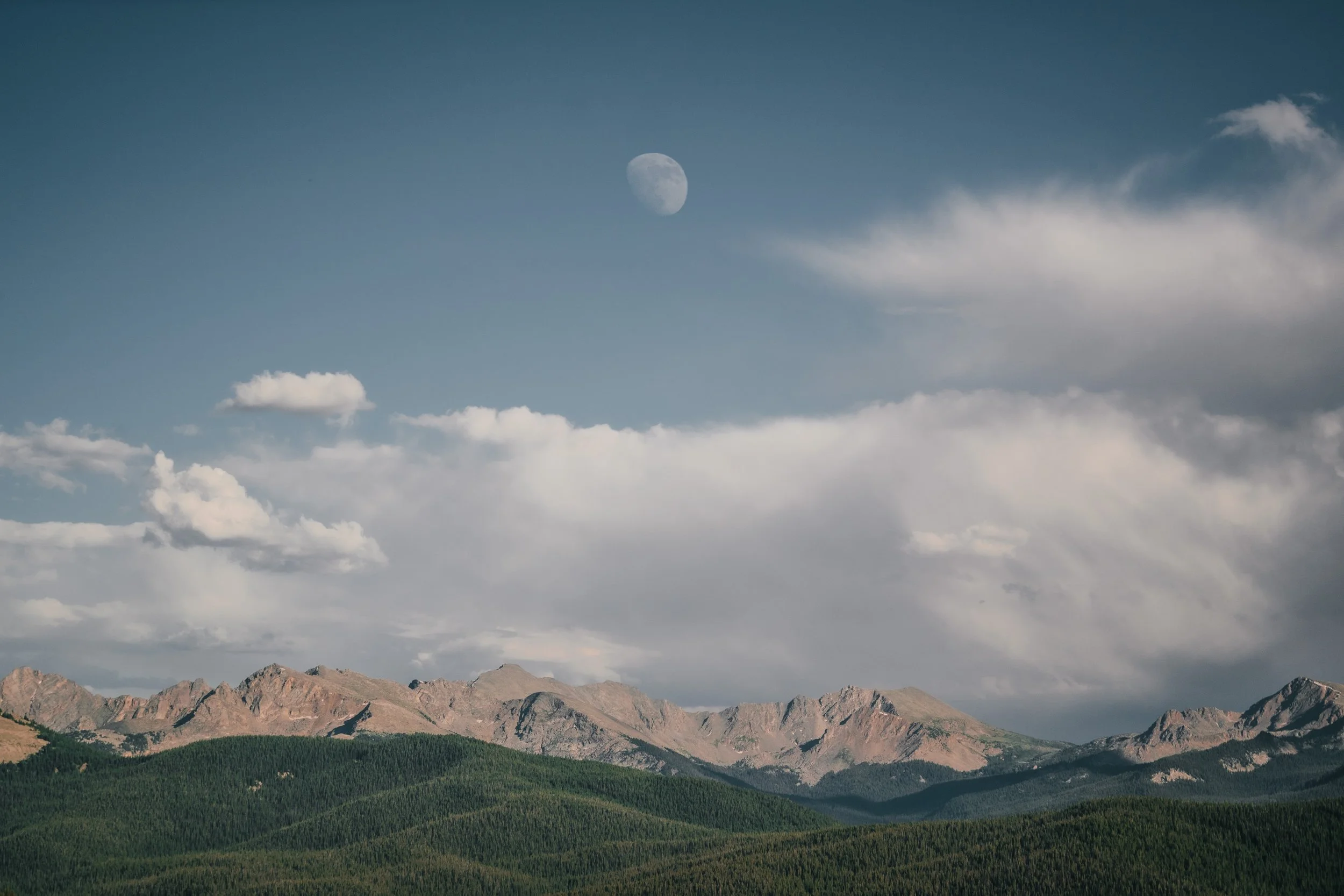 Mountain range with green forests at the base, cloudy sky, and the moon visible in the daytime sky.