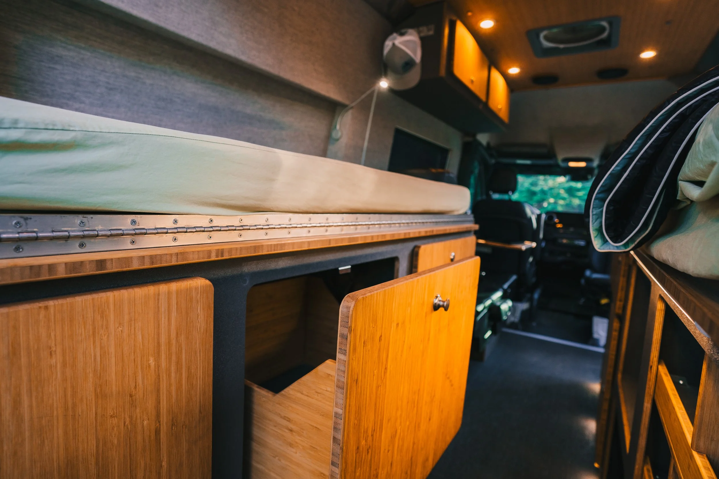 Interior of a camper van with wooden cabinetry, a bed with a green mattress, and a driving area visible in the background.