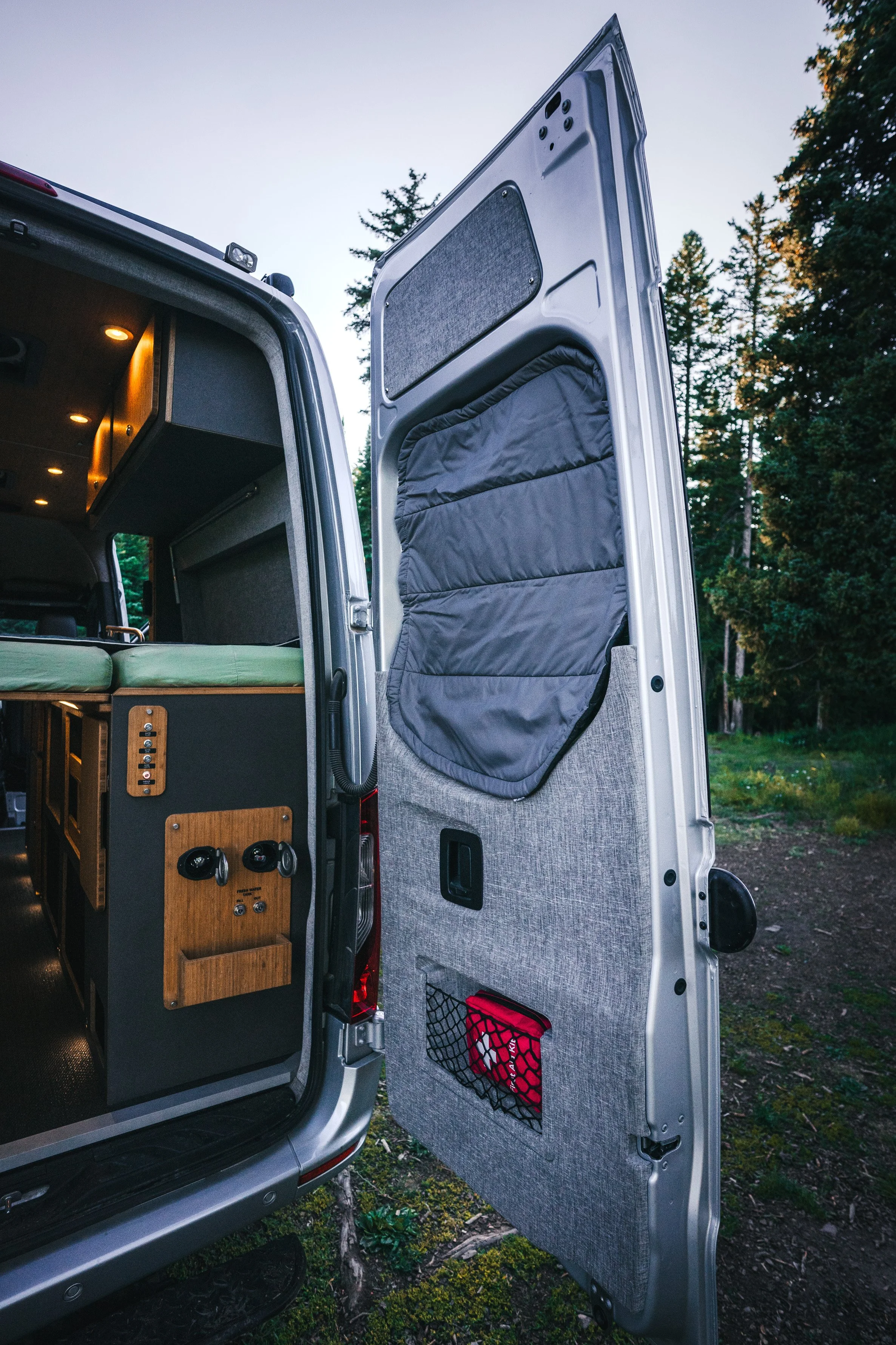 Open van door showing interior with bed, wood panel, and storage compartments on a forested camping site.