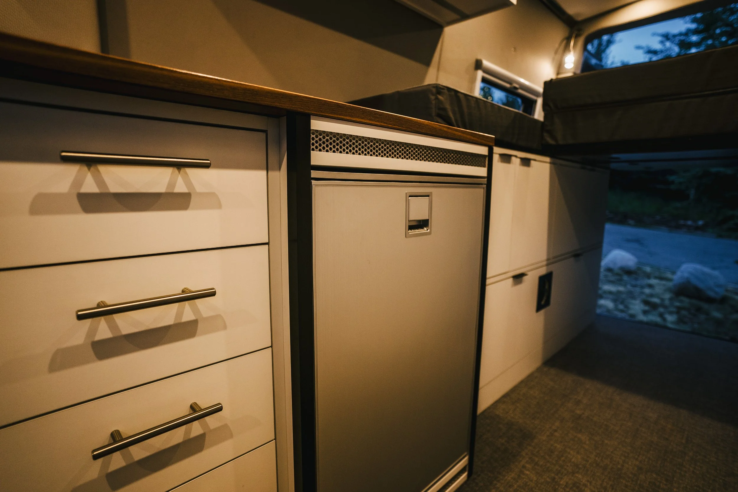 Close-up of a vintage-style kitchen counter with multiple drawers and a mini fridge, in an RV interior during evening.