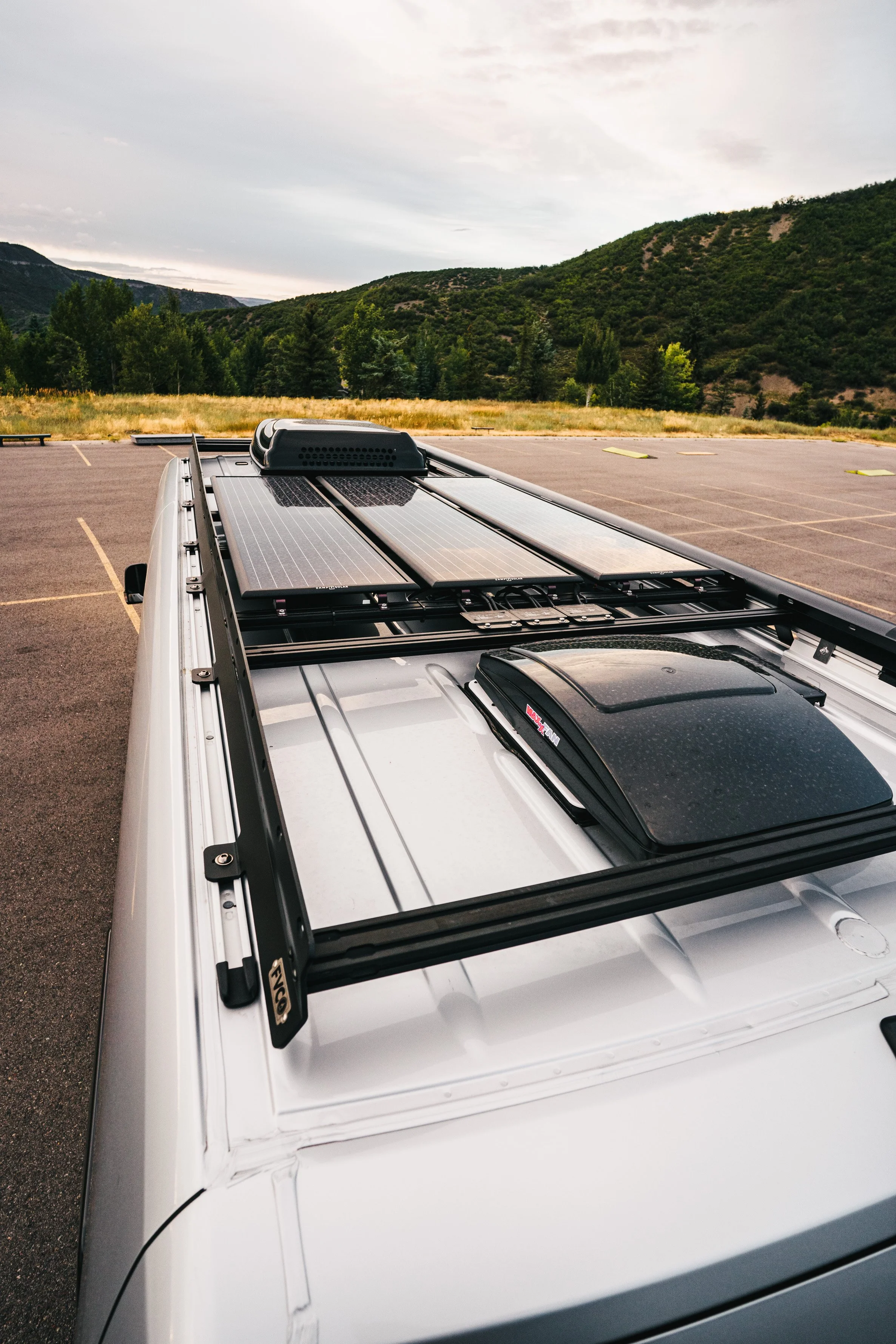 Top view of vehicle roof with solar panels, roof rack, and roof vent, in an empty parking lot surrounded by mountains and trees.