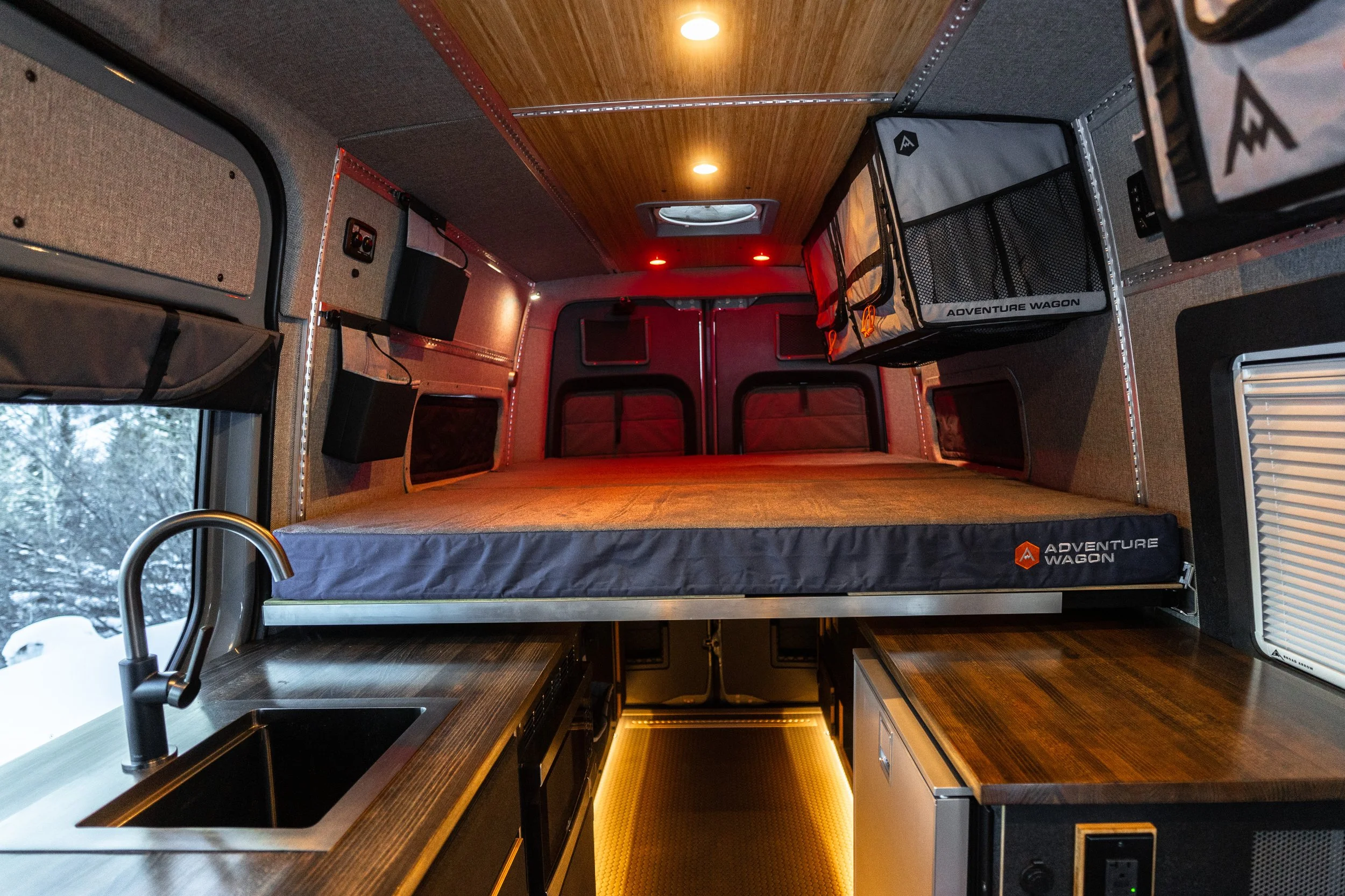 Interior of a camper van with a bed, kitchen sink, and storage compartments, view from the front looking towards the back with snow outside the window.