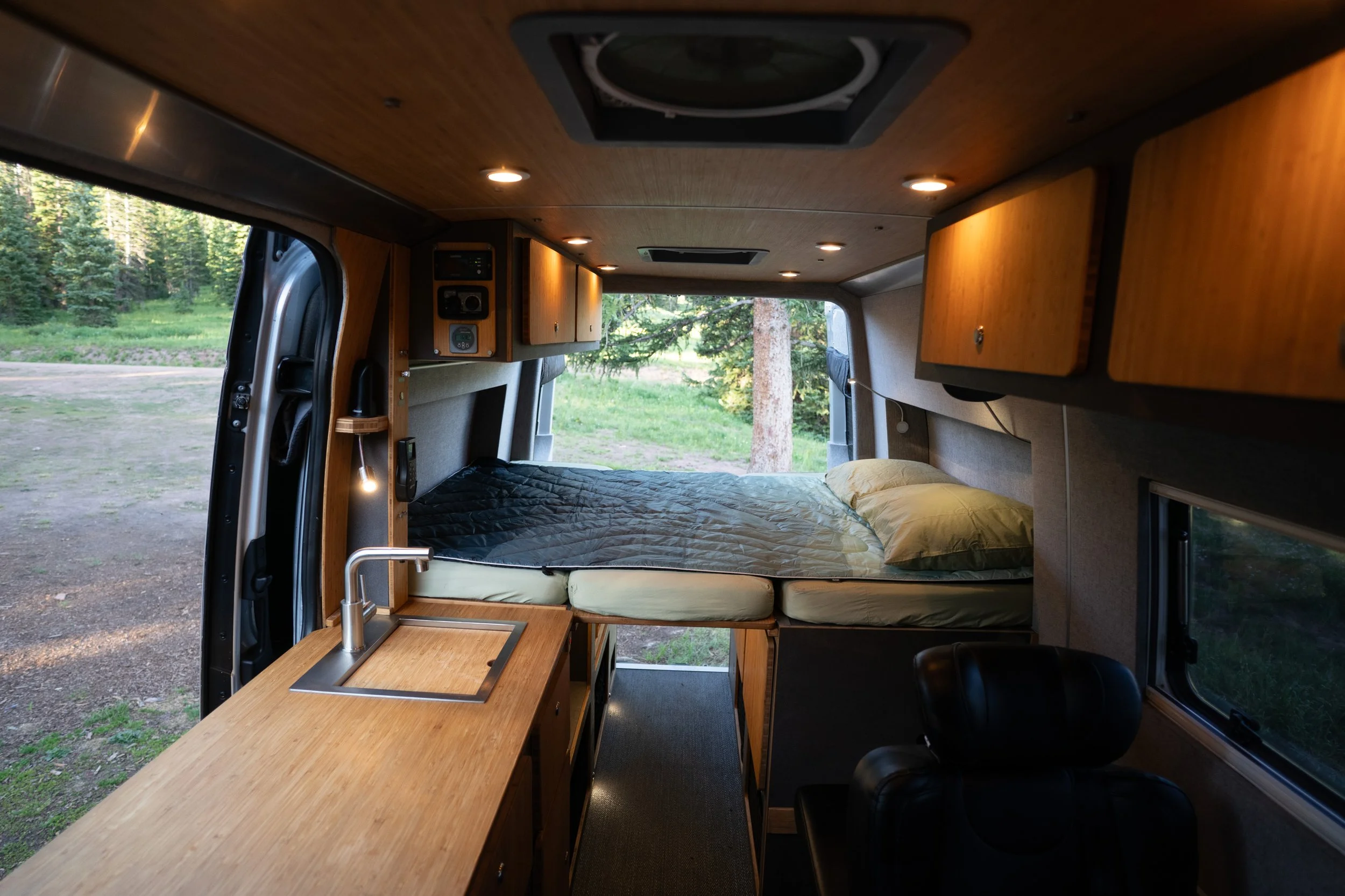 Inside a camper van with a bed, wooden cabinets, a small kitchen with a sink, and a window showing an outdoor green landscape.