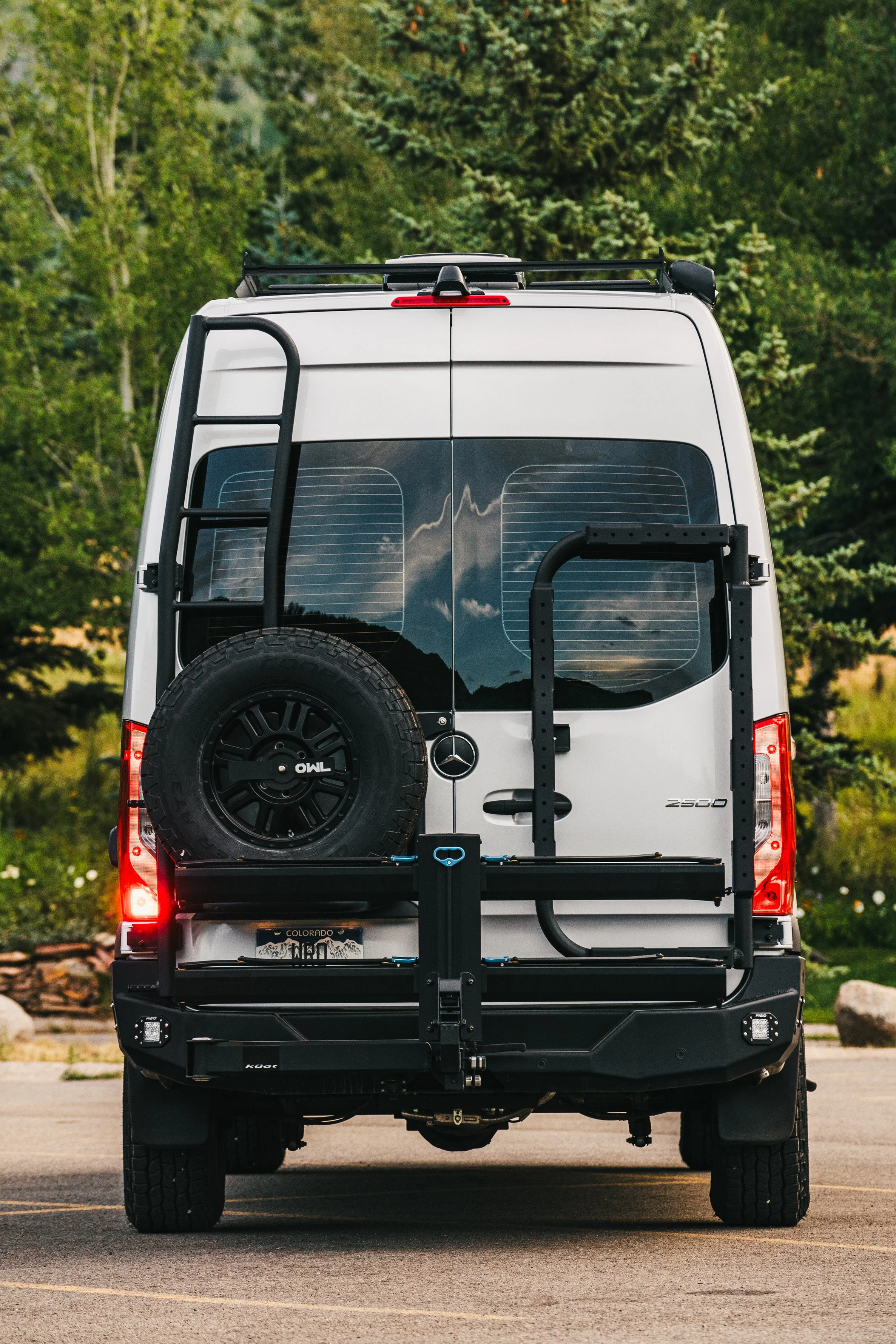 Silver Mercedes-Benz Sprinter van with a spare tire mounted on the back, parked outdoors with trees in the background.