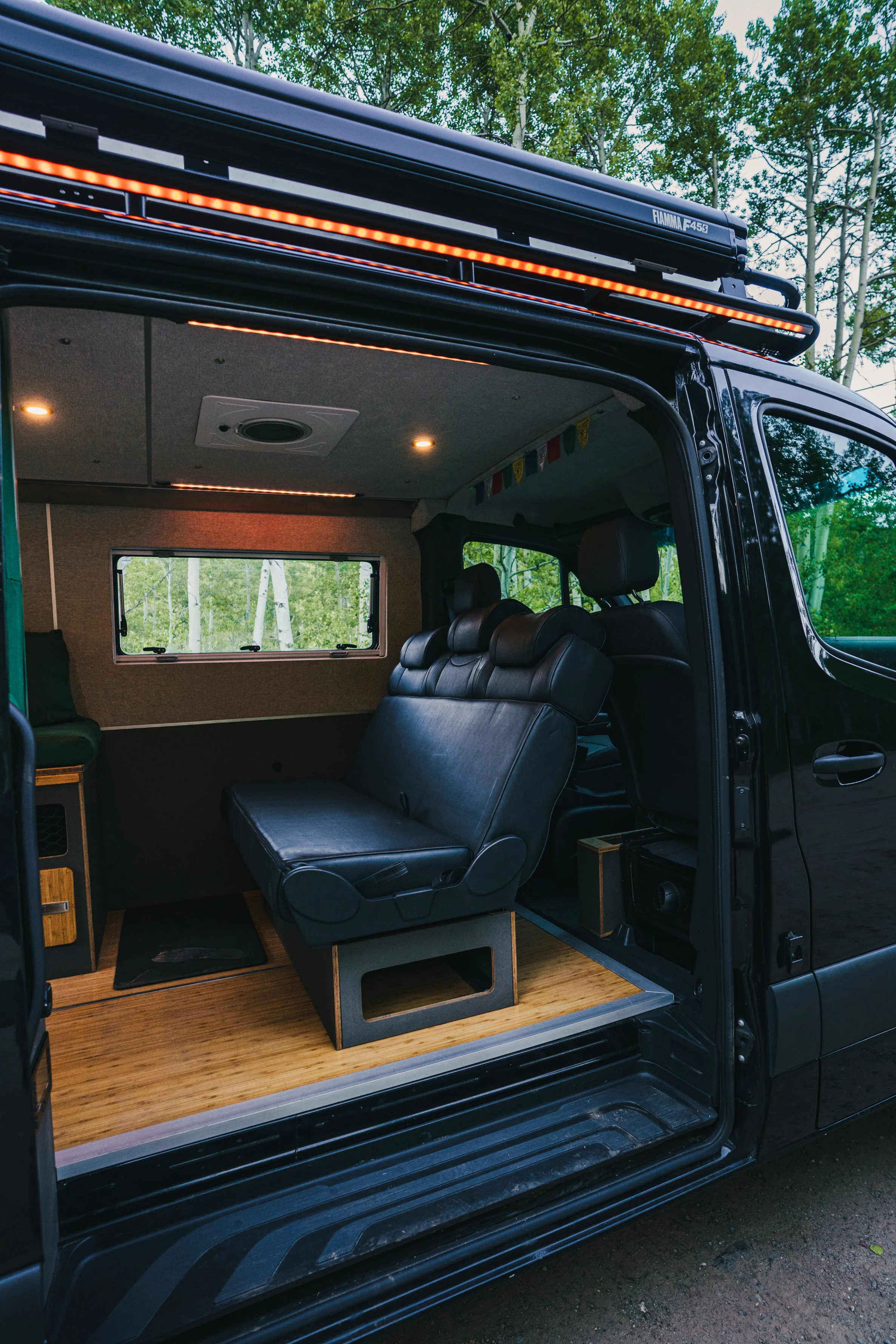 Interior of a converted van with black leather seats, wooden flooring, a window, and ceiling lighting, designed for camping or travel.