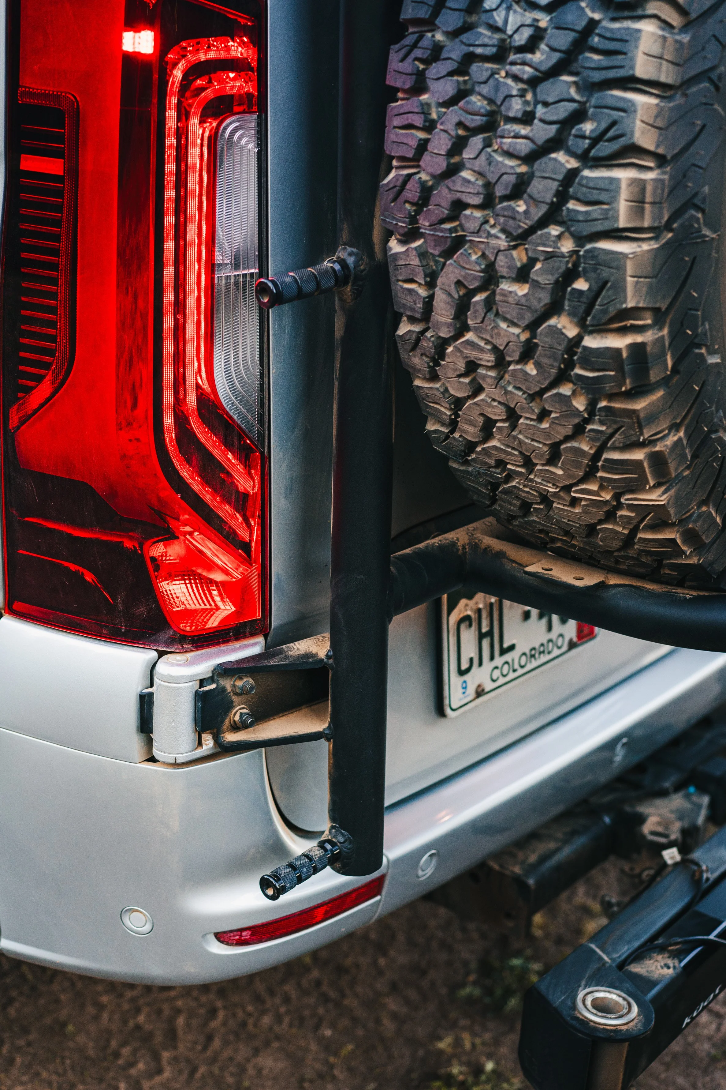 Close-up of the rear of a silver vehicle, showing a large spare tire mounted on a rack, tail light, and Colorado license plate.