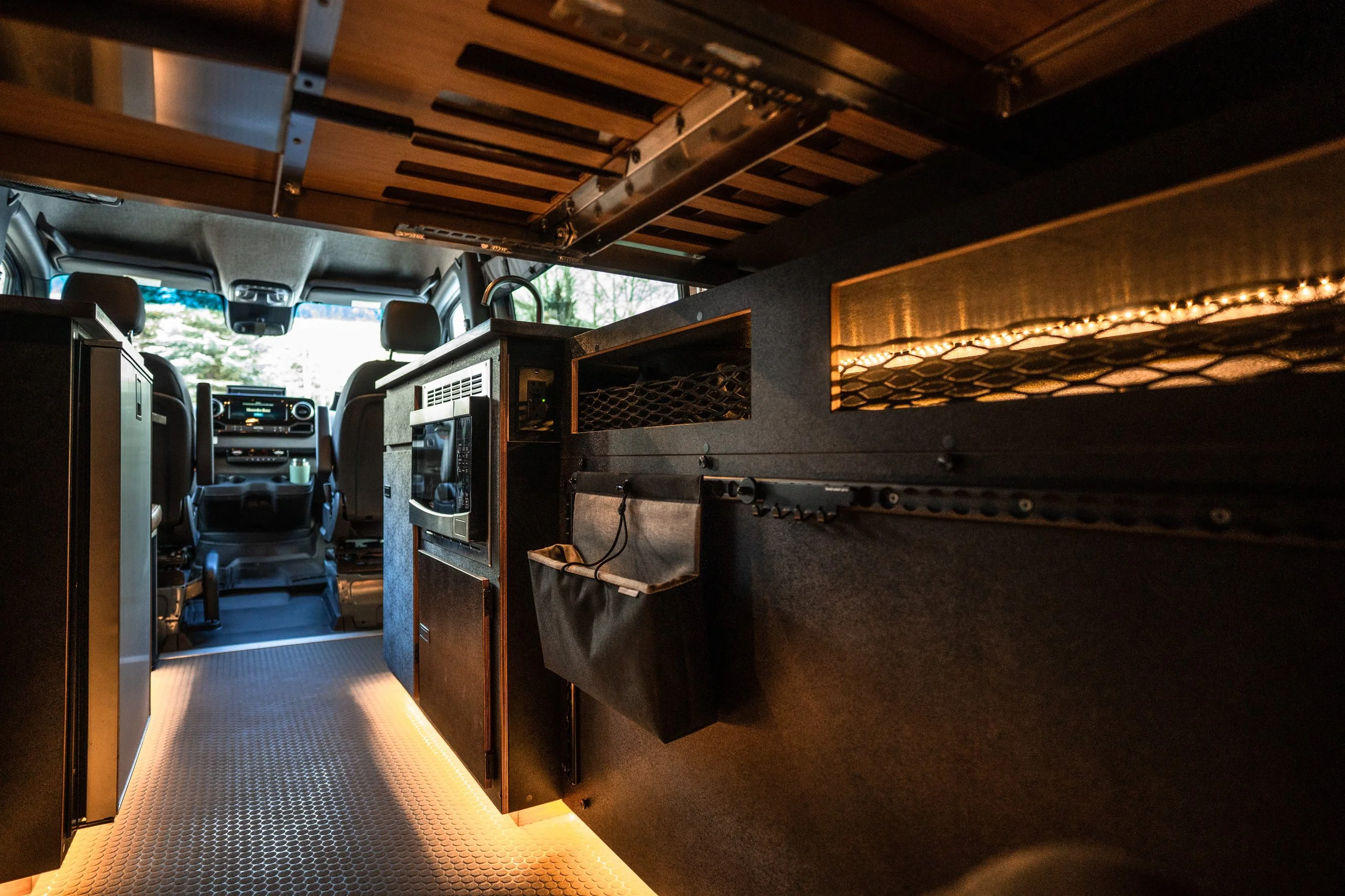 Interior of an RV or camper, showing the driver's cabin at the front and a kitchenette area with a microwave, storage compartments, and under-cabinet lighting.