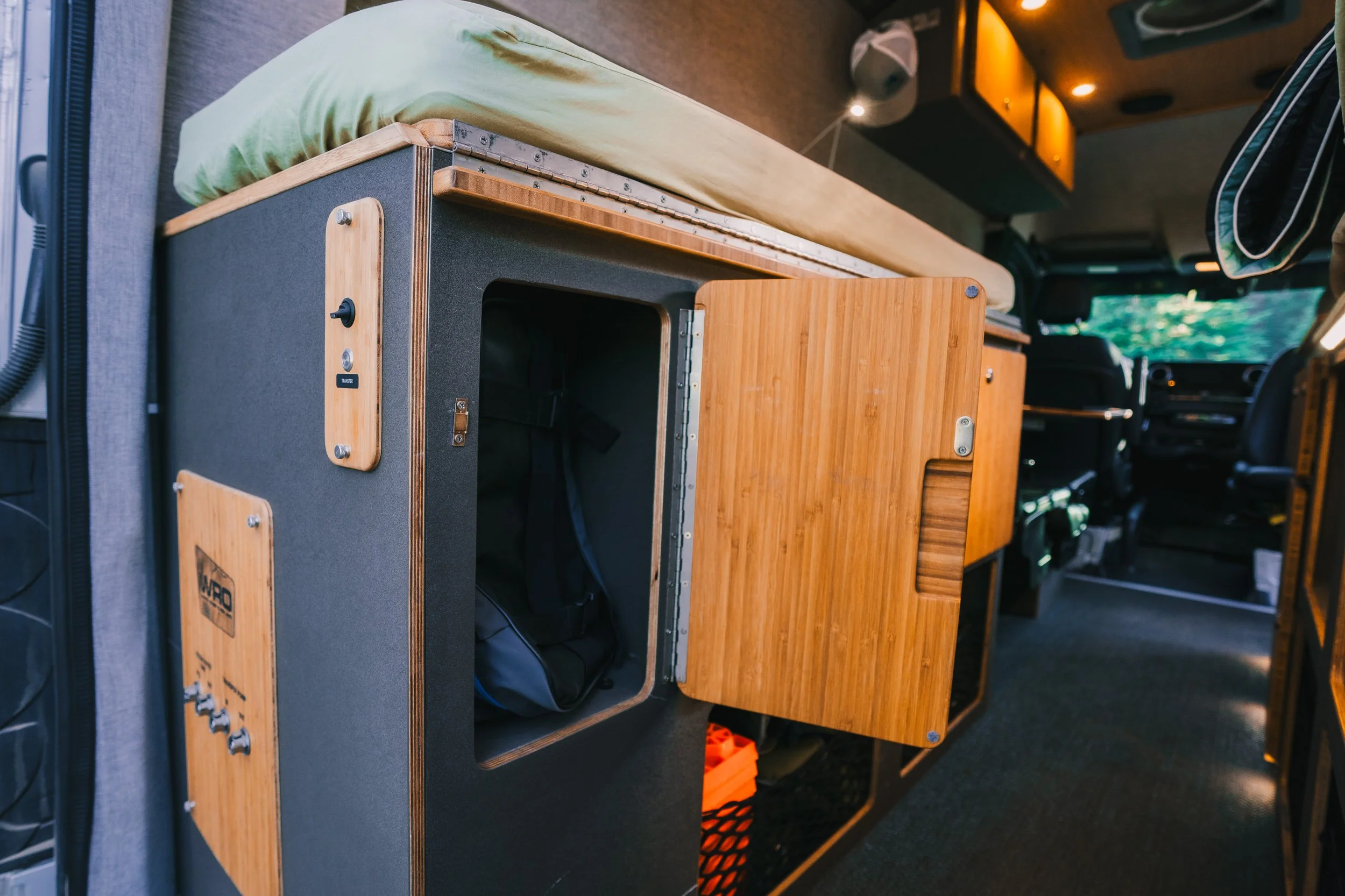 Interior of a converted camper van showing a bed with a pillow and mattress, and a small storage compartment with a wooden door, along with a view of the front vehicle dashboard and windshield.