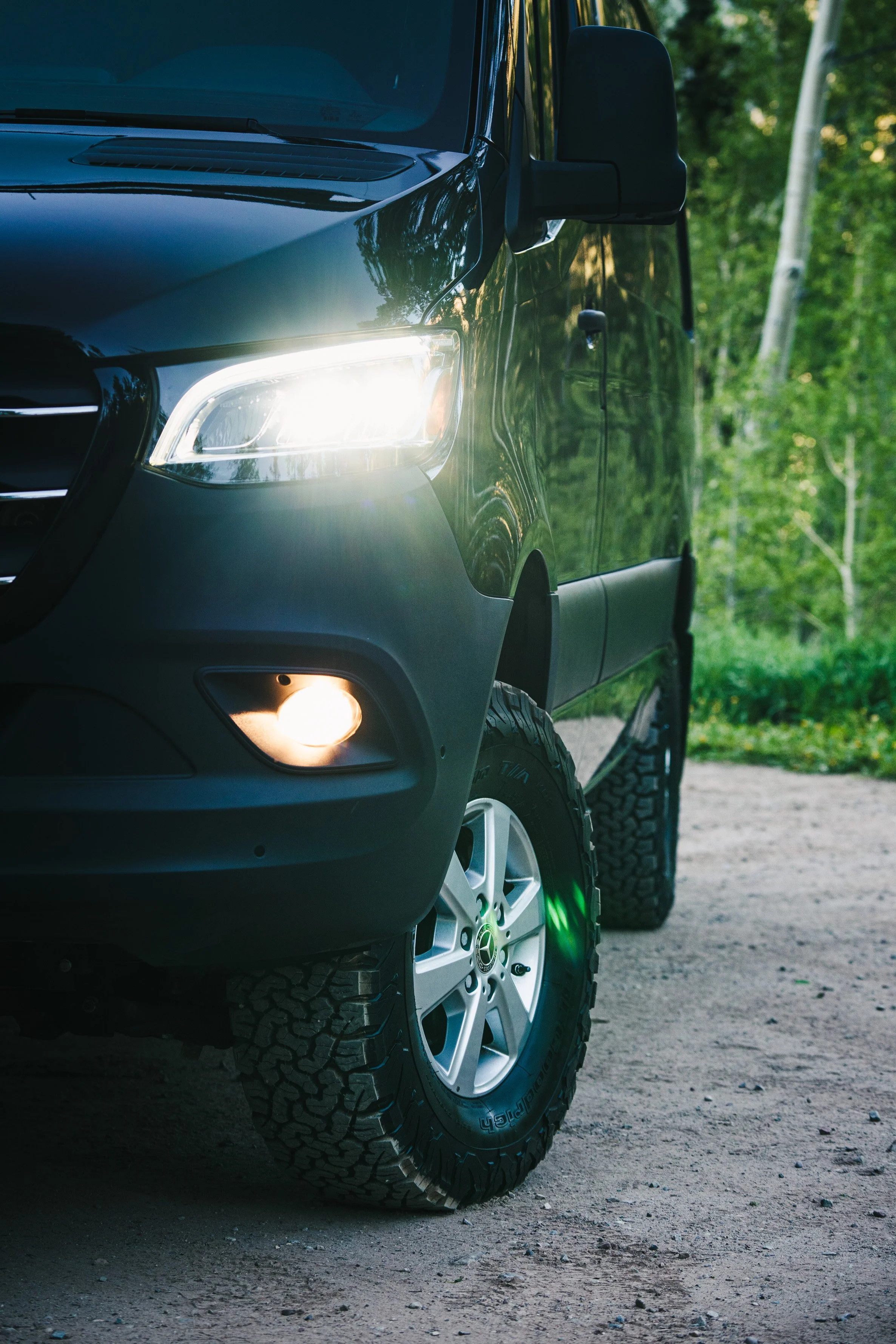 Close-up of the front side of a black SUV with its headlights and fog lights on, parked on a dirt road with green trees in the background.