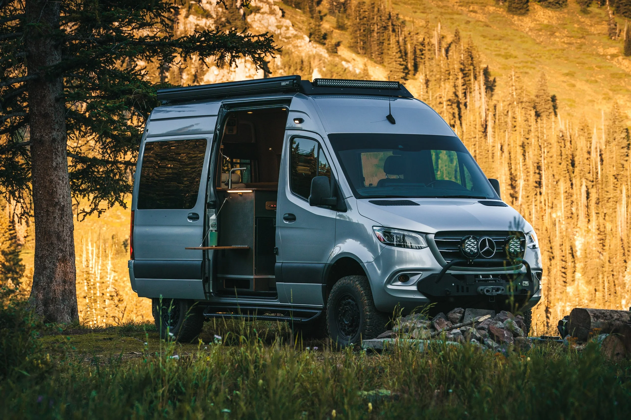 Mercedes-Benz camper van parked in a forest with trees and mountains in the background, with the side door open revealing the interior.