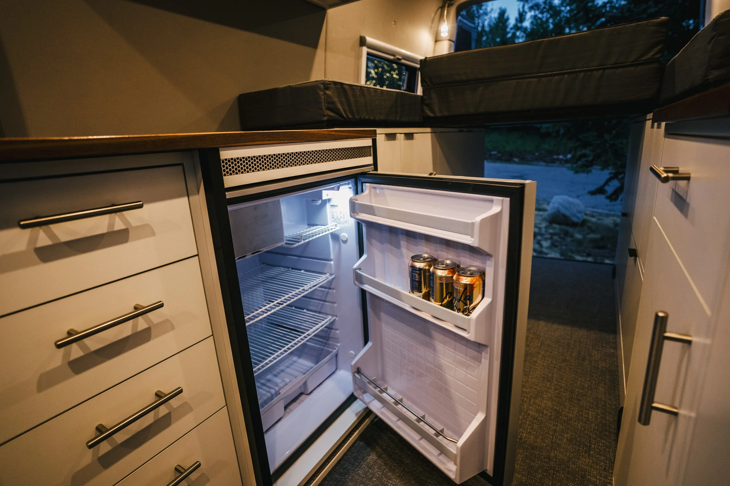 Open small refrigerator in a kitchen with a few cans of soda inside, surrounded by white cabinets and drawers, with a window showing an outdoor scene at dusk.