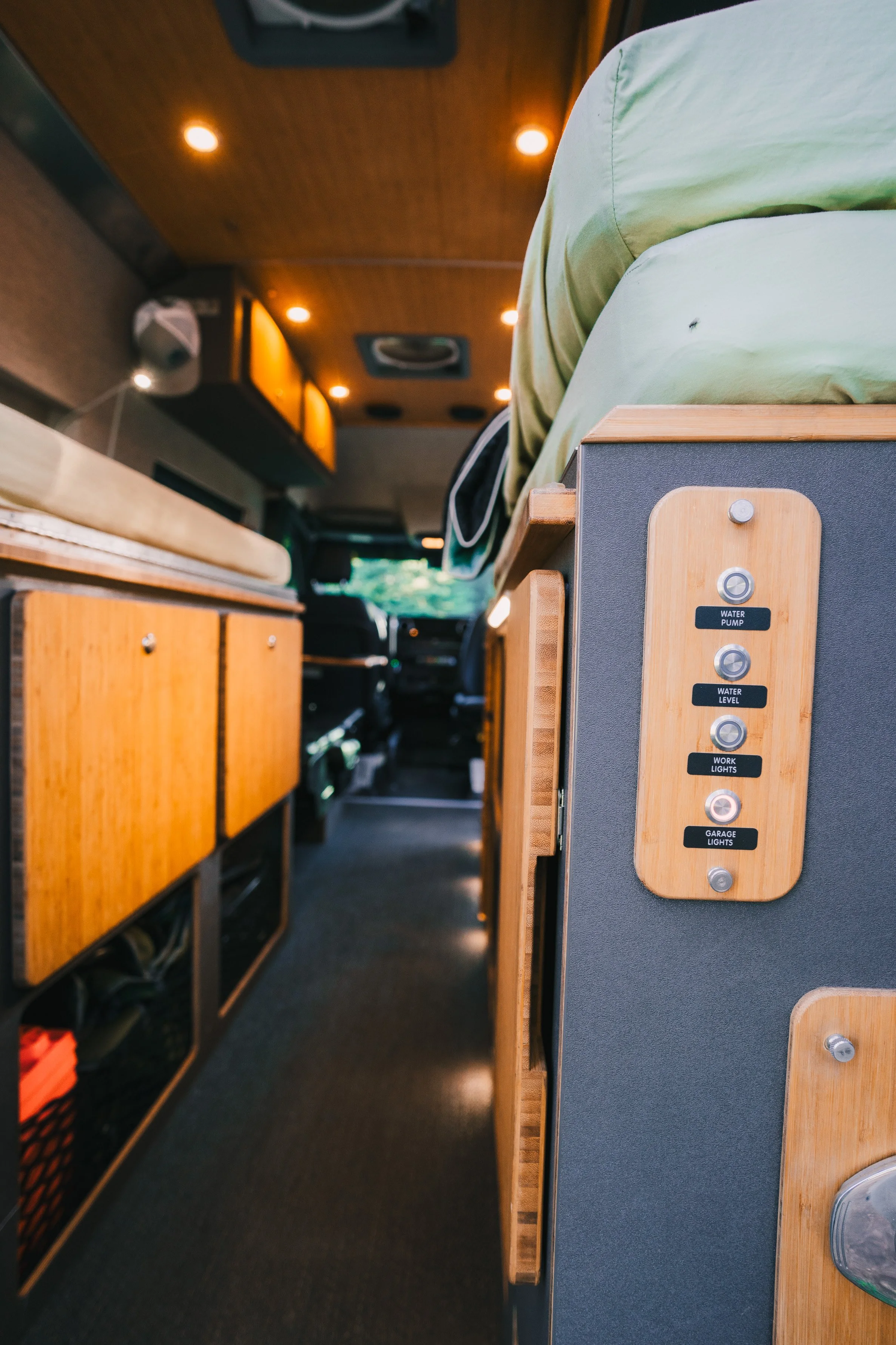Interior of a camper van with wooden cabinets, a person sitting on a bed, and a control panel with labels for water pump, water level, work lights, and garage lights.