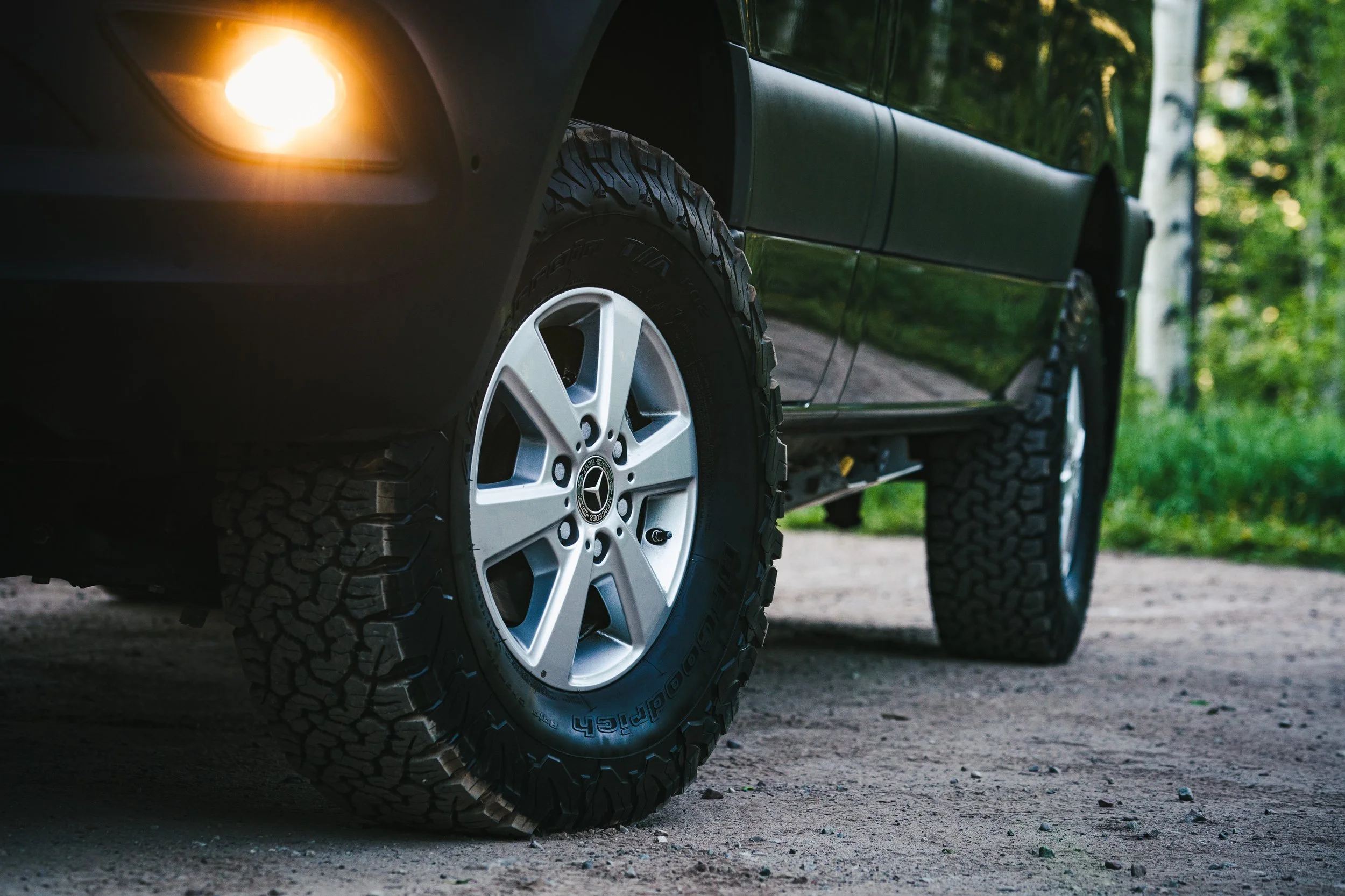 Close-up of a black off-road vehicle with large tires, parked on a dirt road with greenery in the background. The front left tire and part of the vehicle's body are visible, with a vehicle headlight turned on.