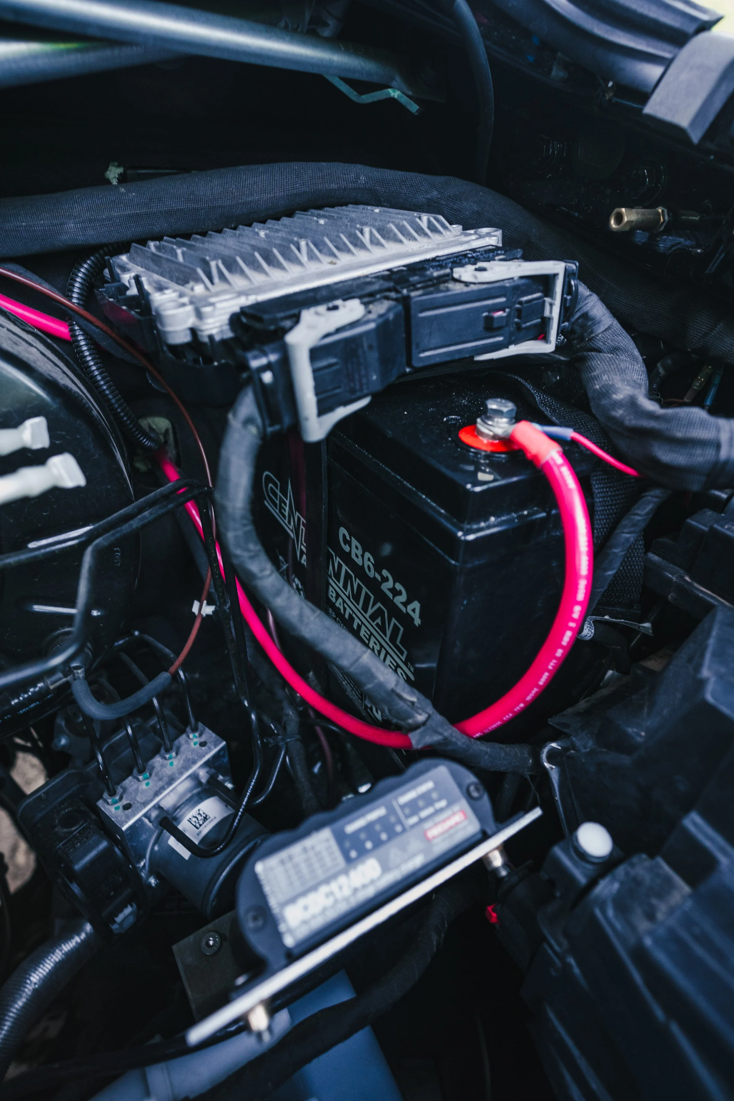 Inside a vehicle engine bay with a car battery and electronic control module, with various cables and wires connected.