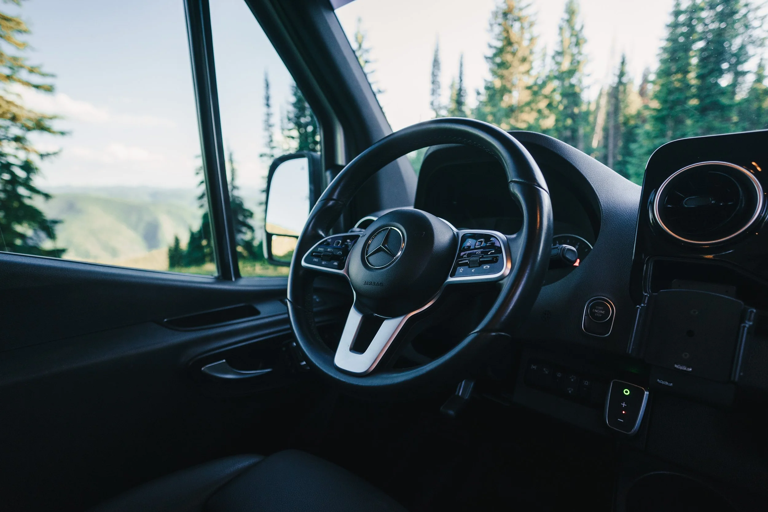 Inside view of a Mercedes-Benz vehicle's dashboard and steering wheel, with a scenic forest visible through the windshield.