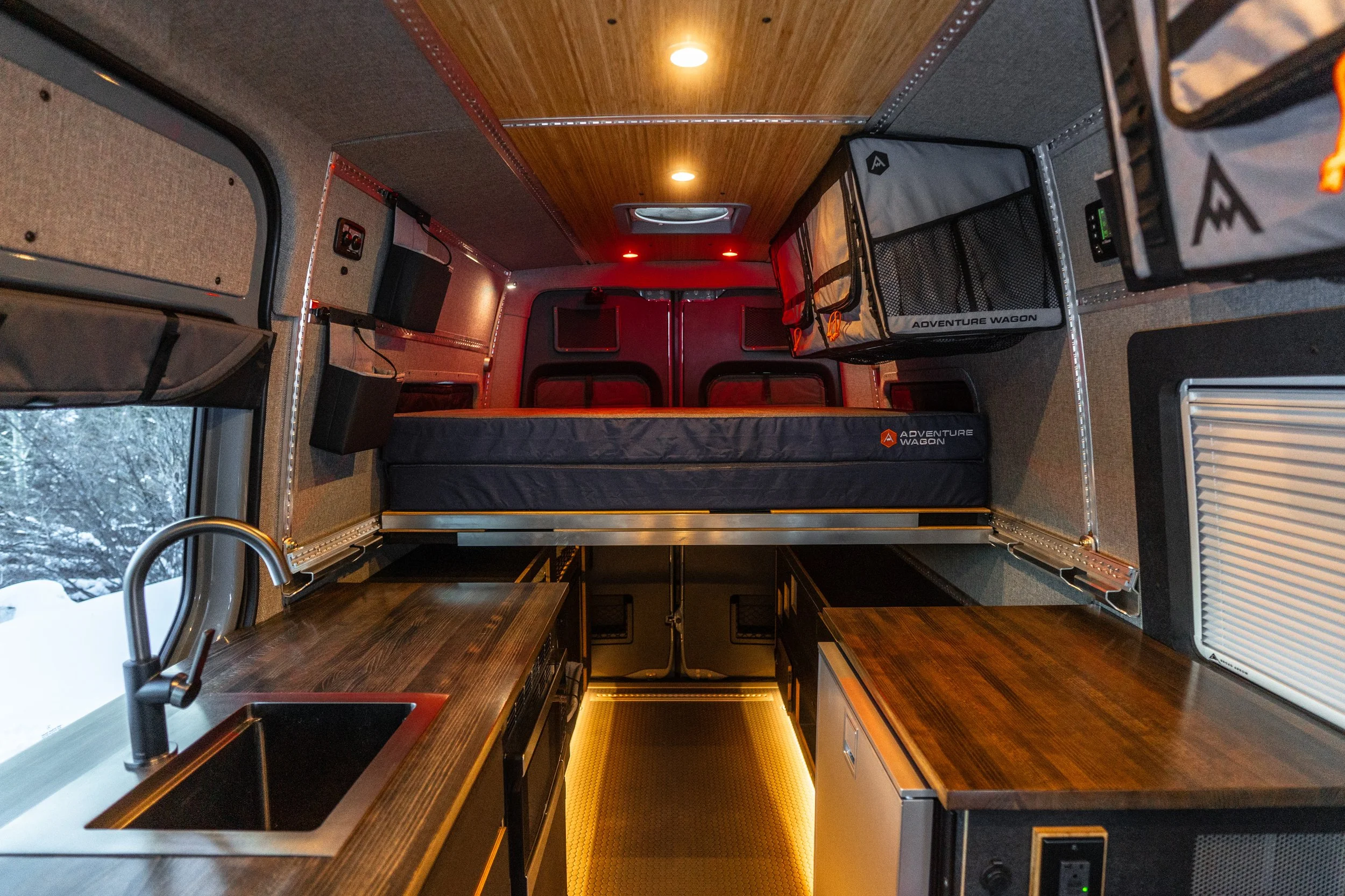 Interior of a custom camper van with a kitchenette, wooden countertops, a small sink, a stove, and a fold-up sleeping platform with Adventure Wagon gear, illuminated by ceiling lights.