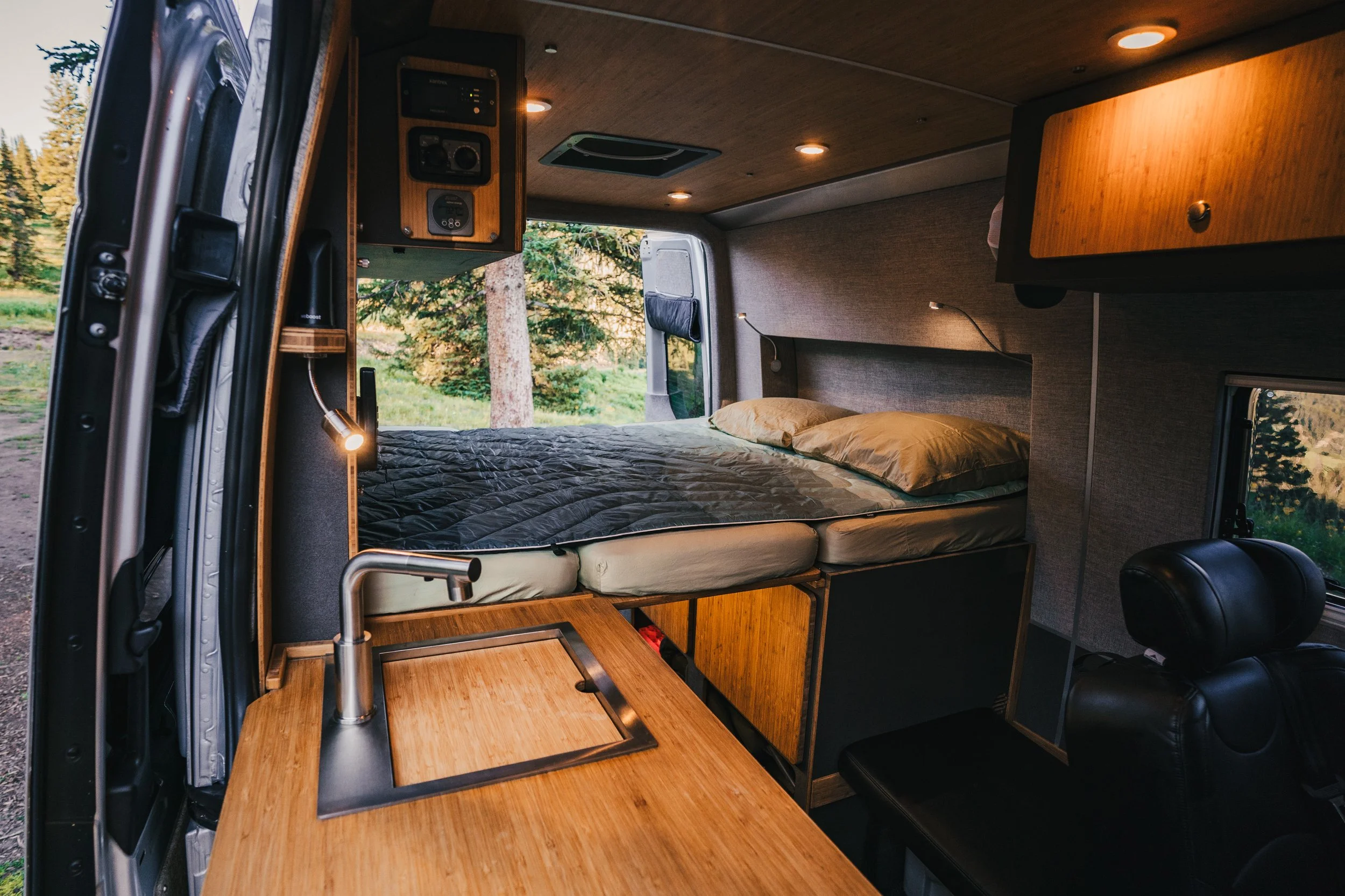 Interior of a camper van with a bed, small kitchen with sink, and seating area, set in a natural outdoor environment.