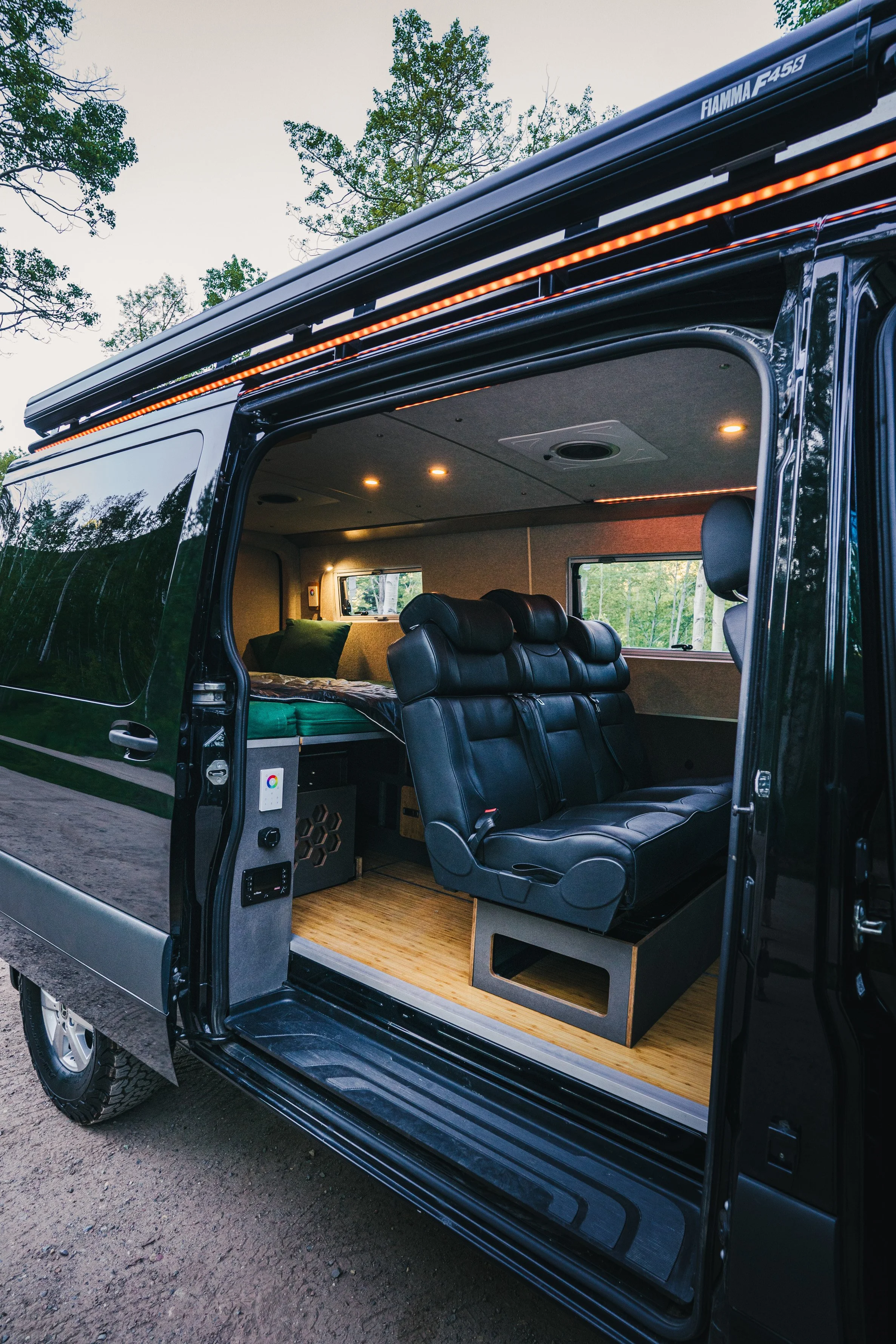 Front side of a converted van with sliding door open showing a bedroom with a bed, black leather seats, and a wood floor, parked outdoors with trees in the background.