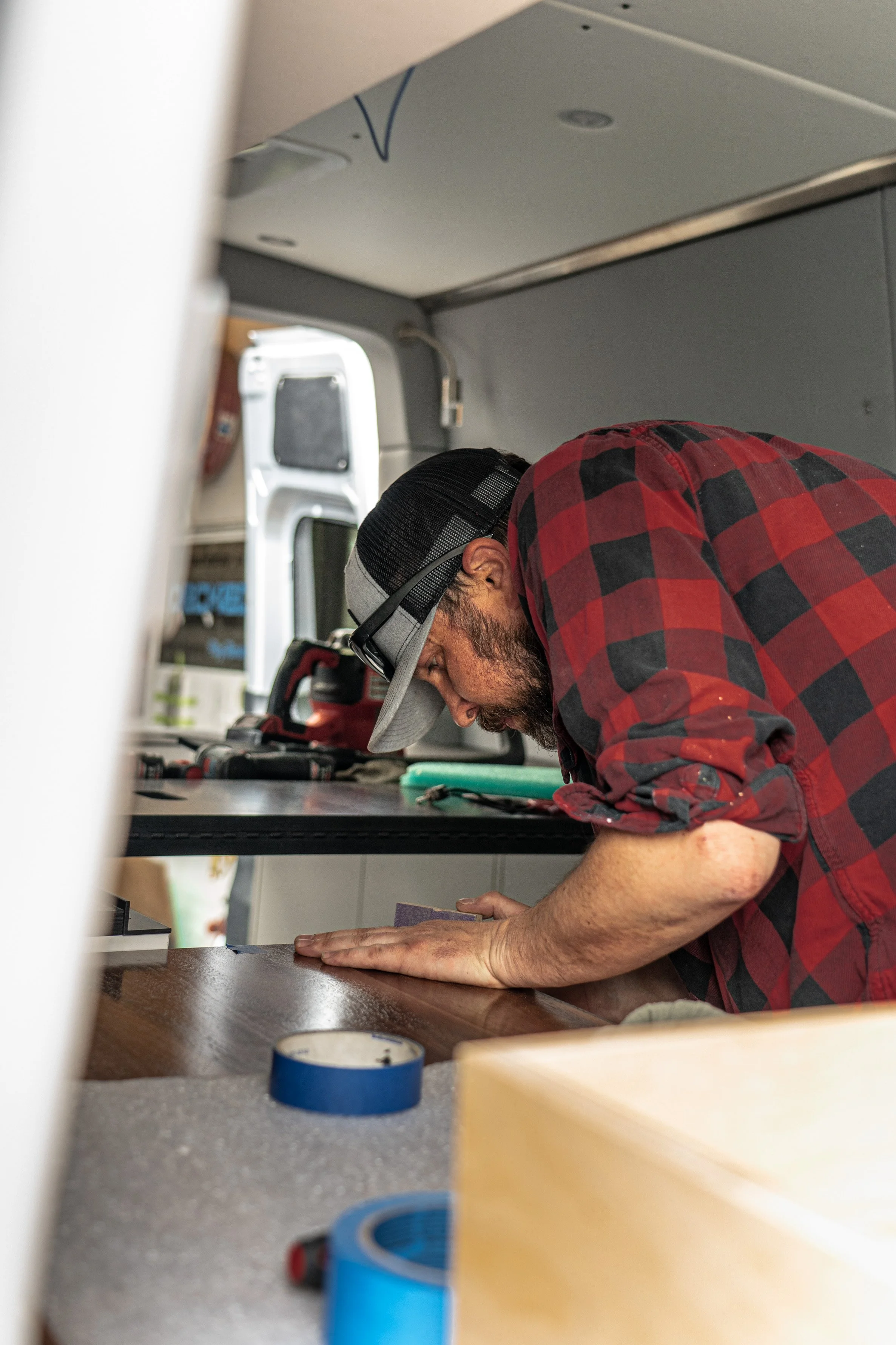 A man wearing a red and black checkered shirt, gray and black cap, and glasses leans over a dark wooden table that has painter's tape and a piece of sandpaper on it, working on a project inside a van, with tools and equipment on the countertop and an open van door in the background.