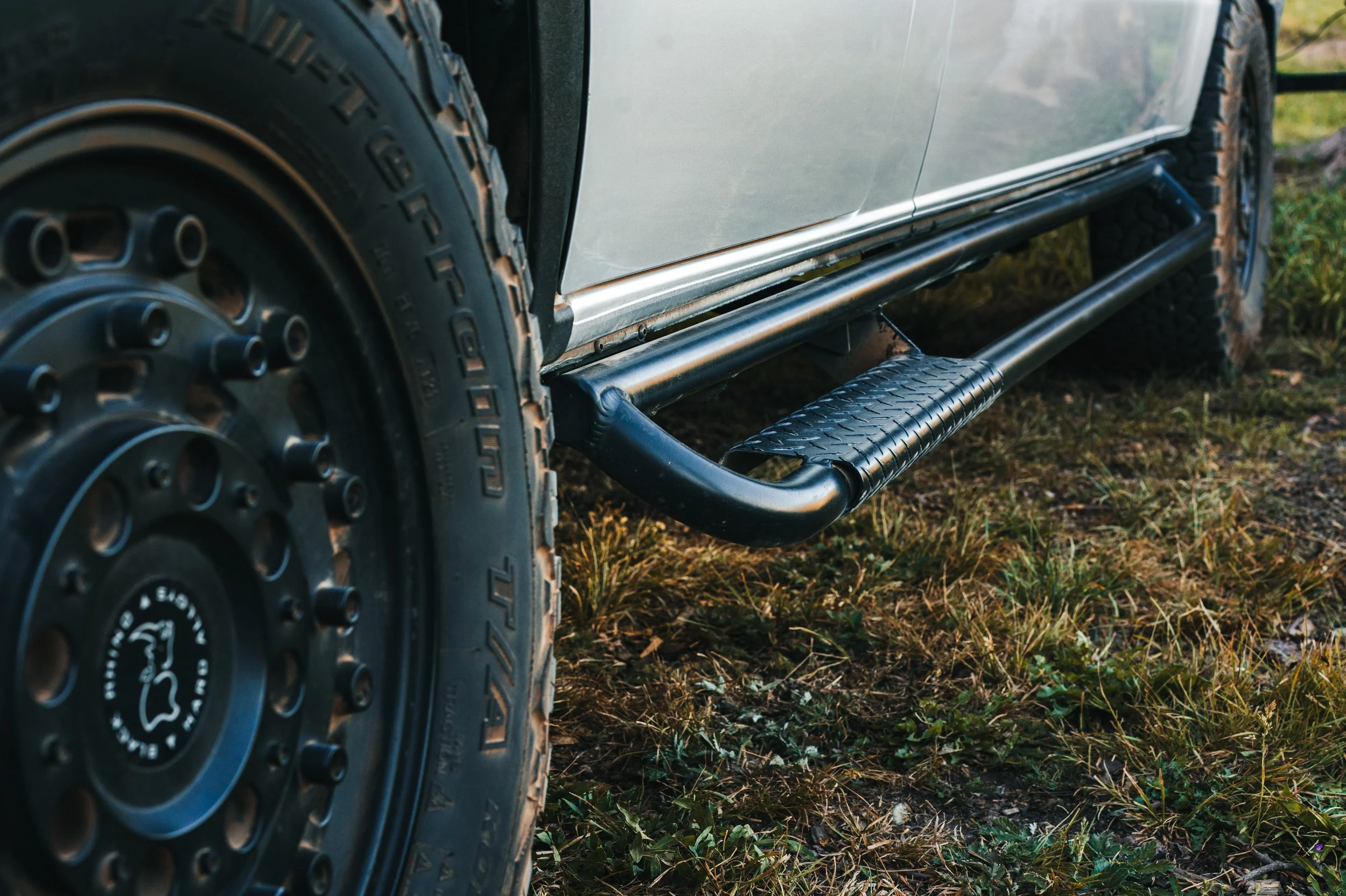 Close-up of a vehicle's tire and running board, with a grassy ground beneath.
