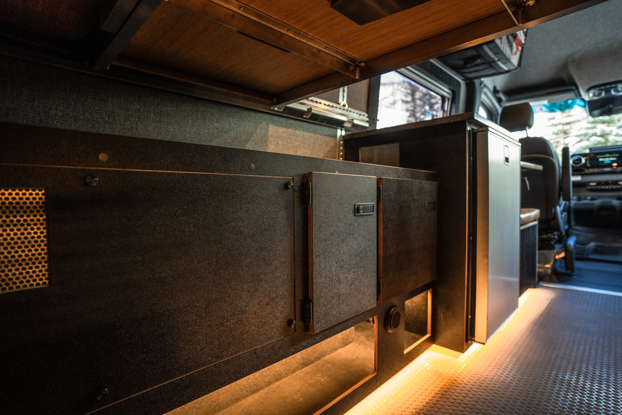 Interior shot of a camper van or bus, showing a black cabinet with a small door, illuminated by yellow lighting, with a window and driver area visible in the background.
