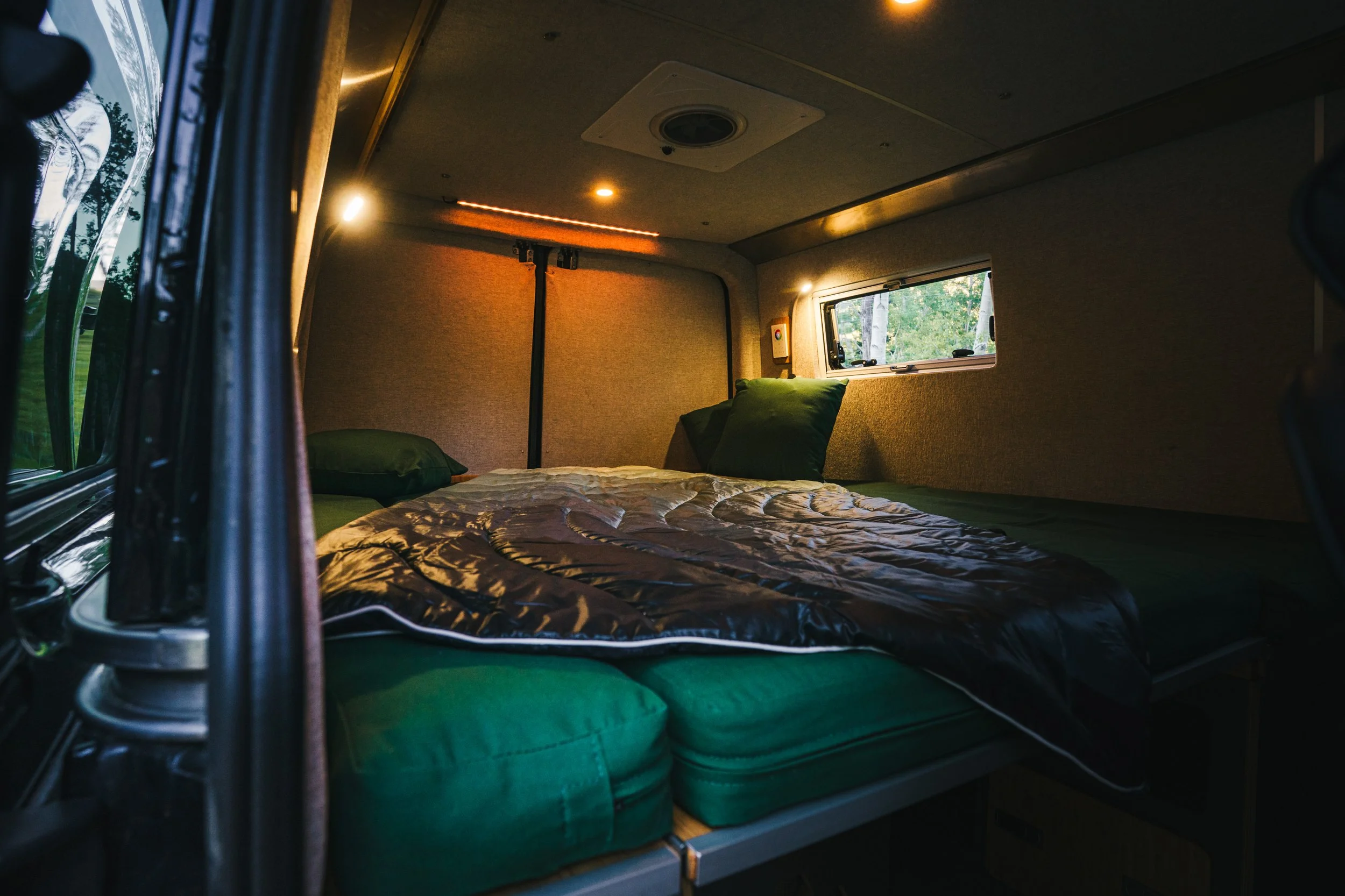 Inside a camper van, a bed with green bedding and pillows is shown, with a small window showing trees outside. The space has warm lighting and beige wall panels.