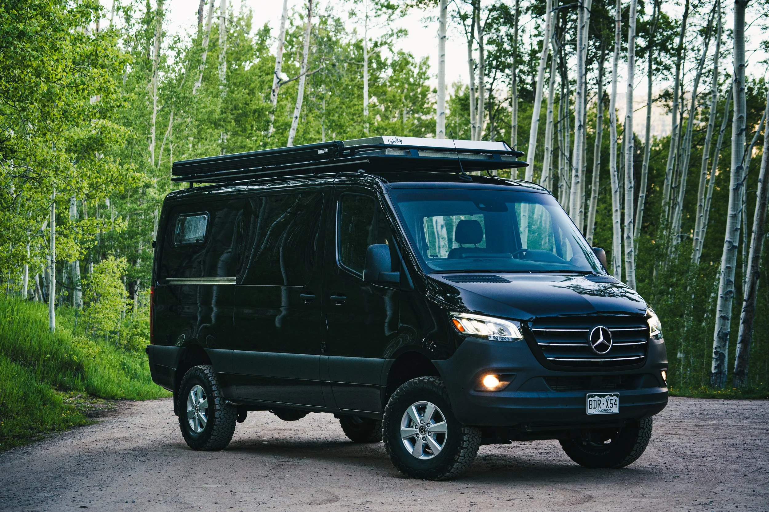 Black Mercedes-Benz camper van parked on a dirt road in a forest with tall green trees in the background.
