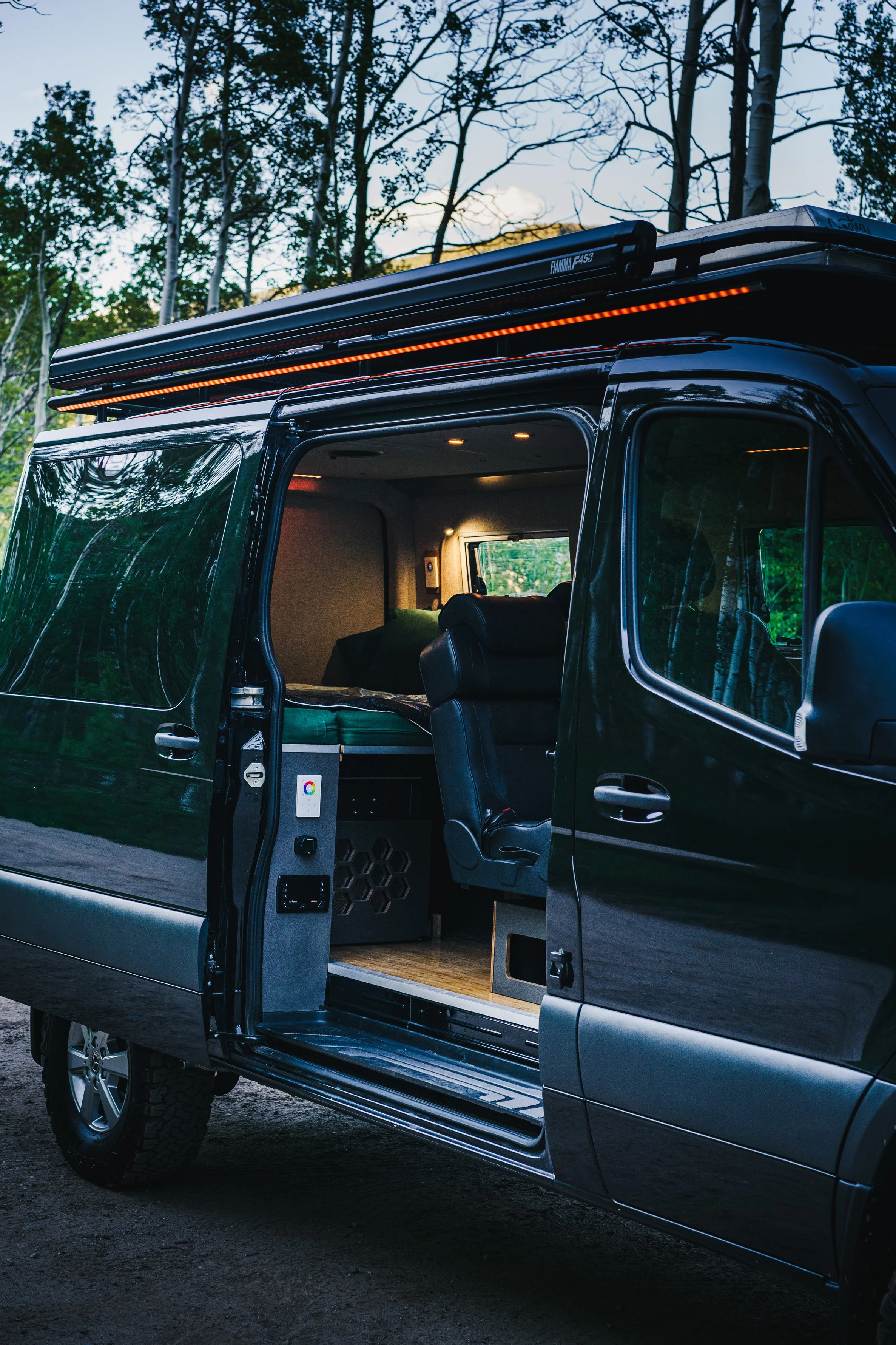 Black van with sliding door open, showing a key, a control panel, and a seating area inside, parked on a dirt path in a wooded area during daytime