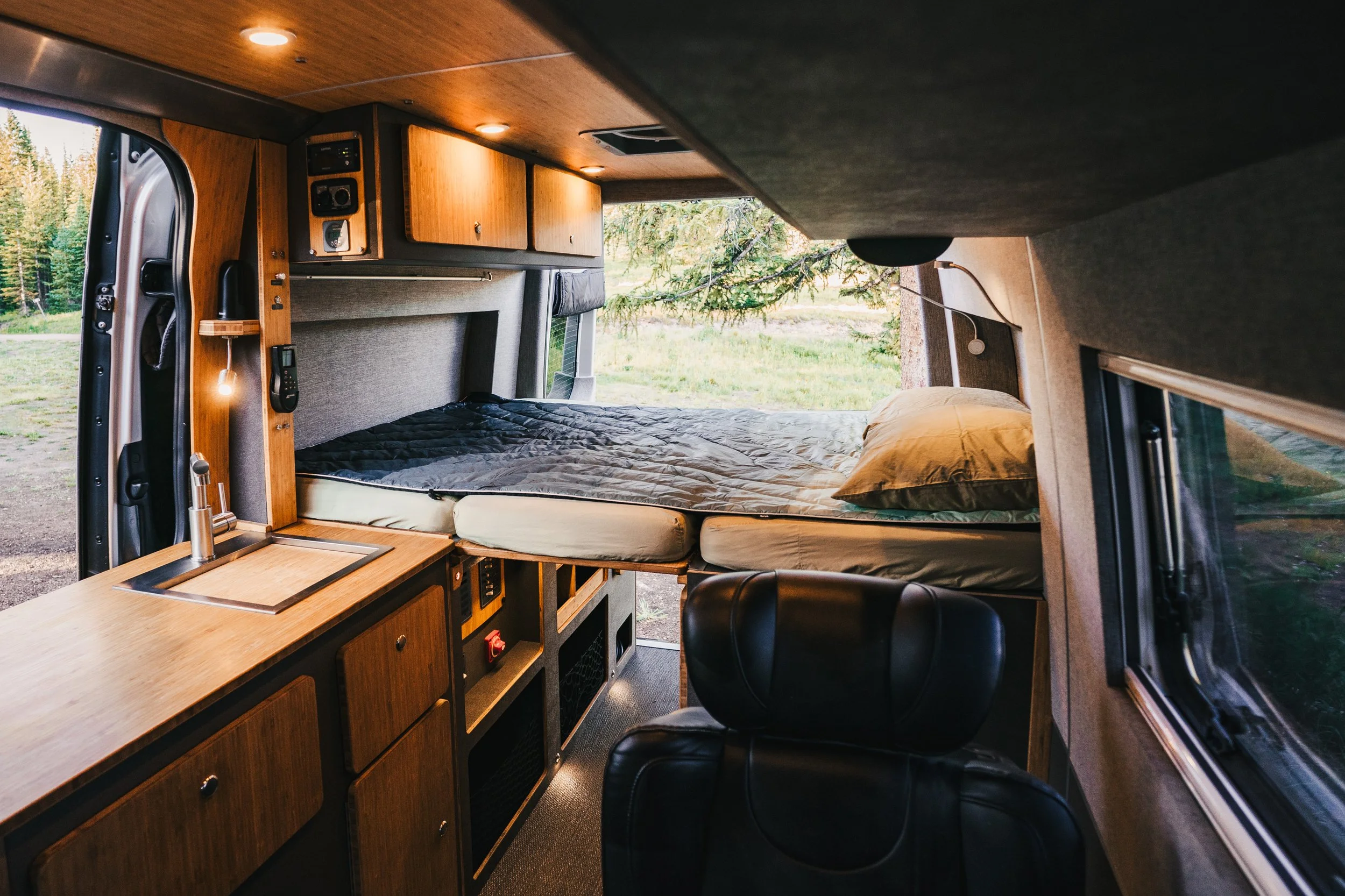 Interior of a camper van with a bed, wooden cabinets, a small sink, and a window showing a forested outdoor scene.