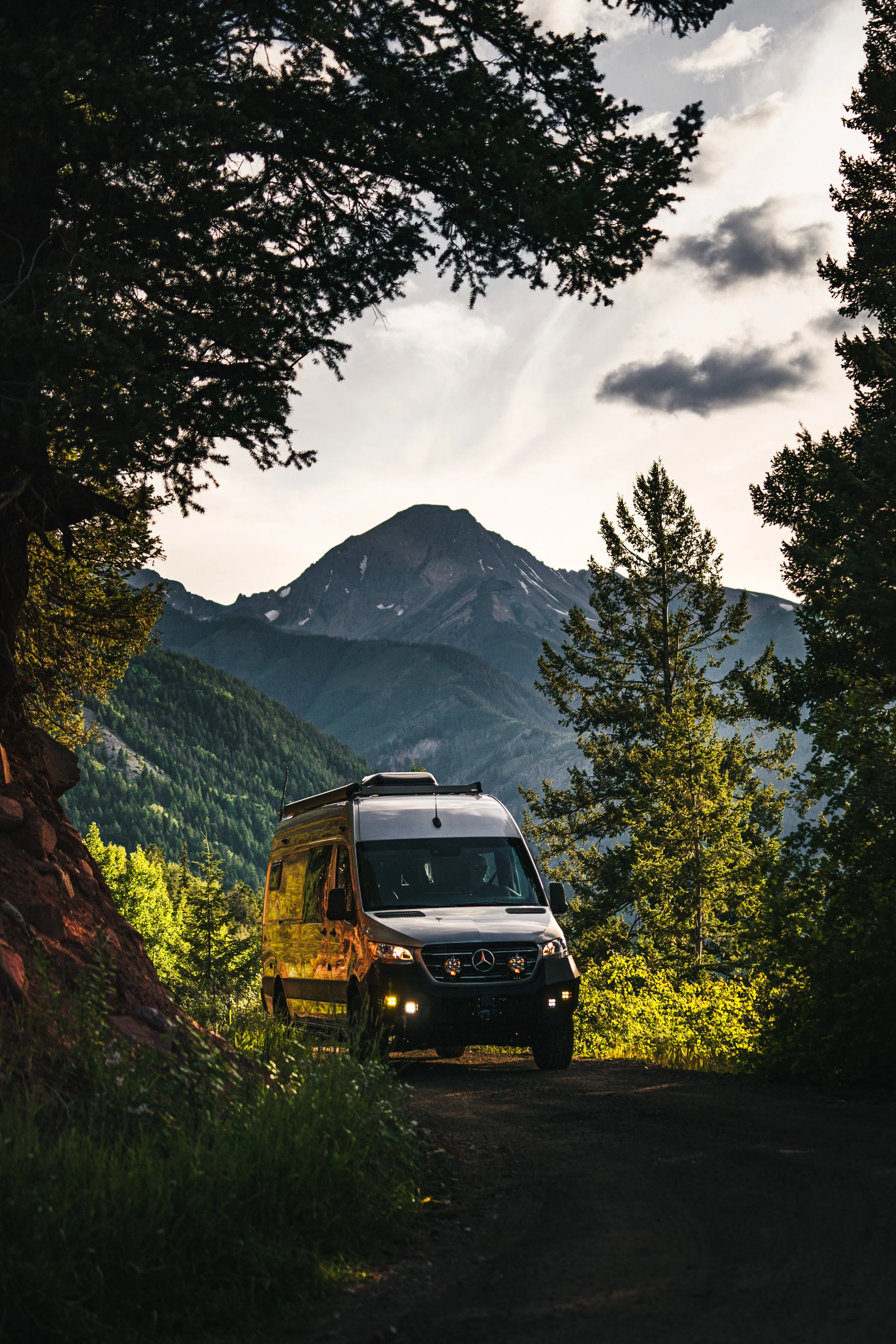 A silver Mercedes-Benz van parked on a dirt trail in a forest, with a mountain in the background and trees framing the scene.