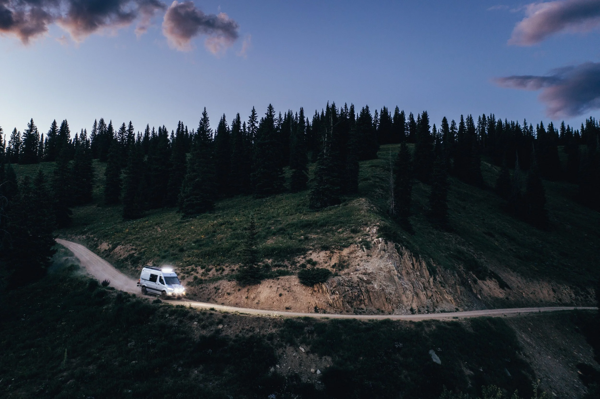 A white van driving on a winding dirt road through a forested hillside during dusk or dawn with a cloudy sky.