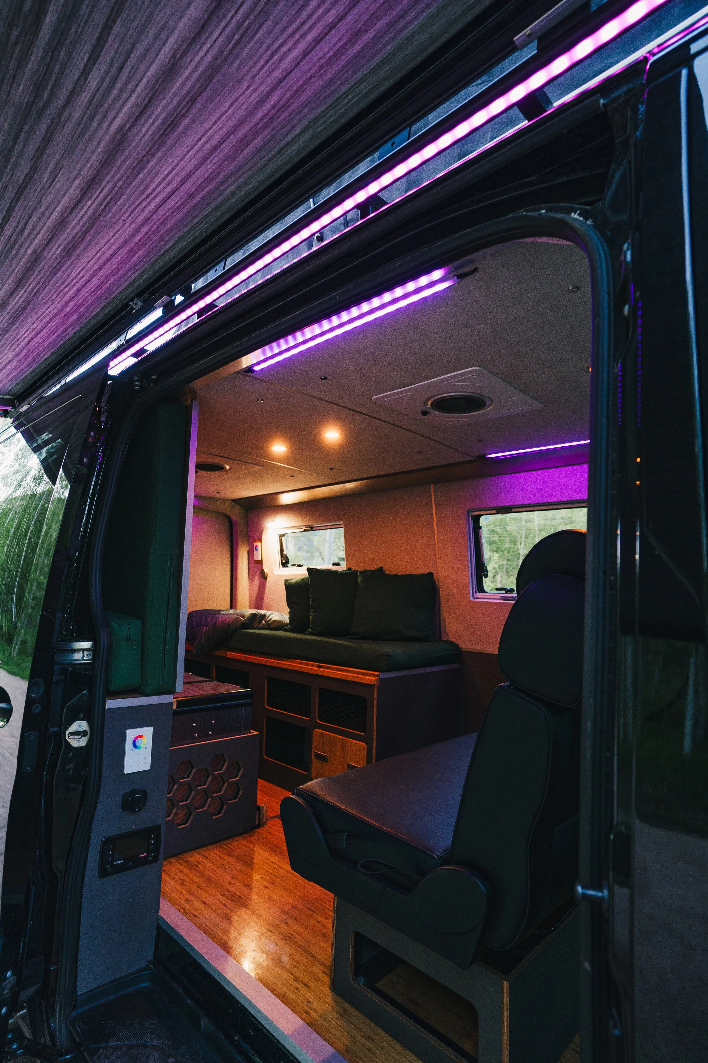Inside a converted van with wooden flooring, black cushioned seats, purple LED lighting, and small windows.