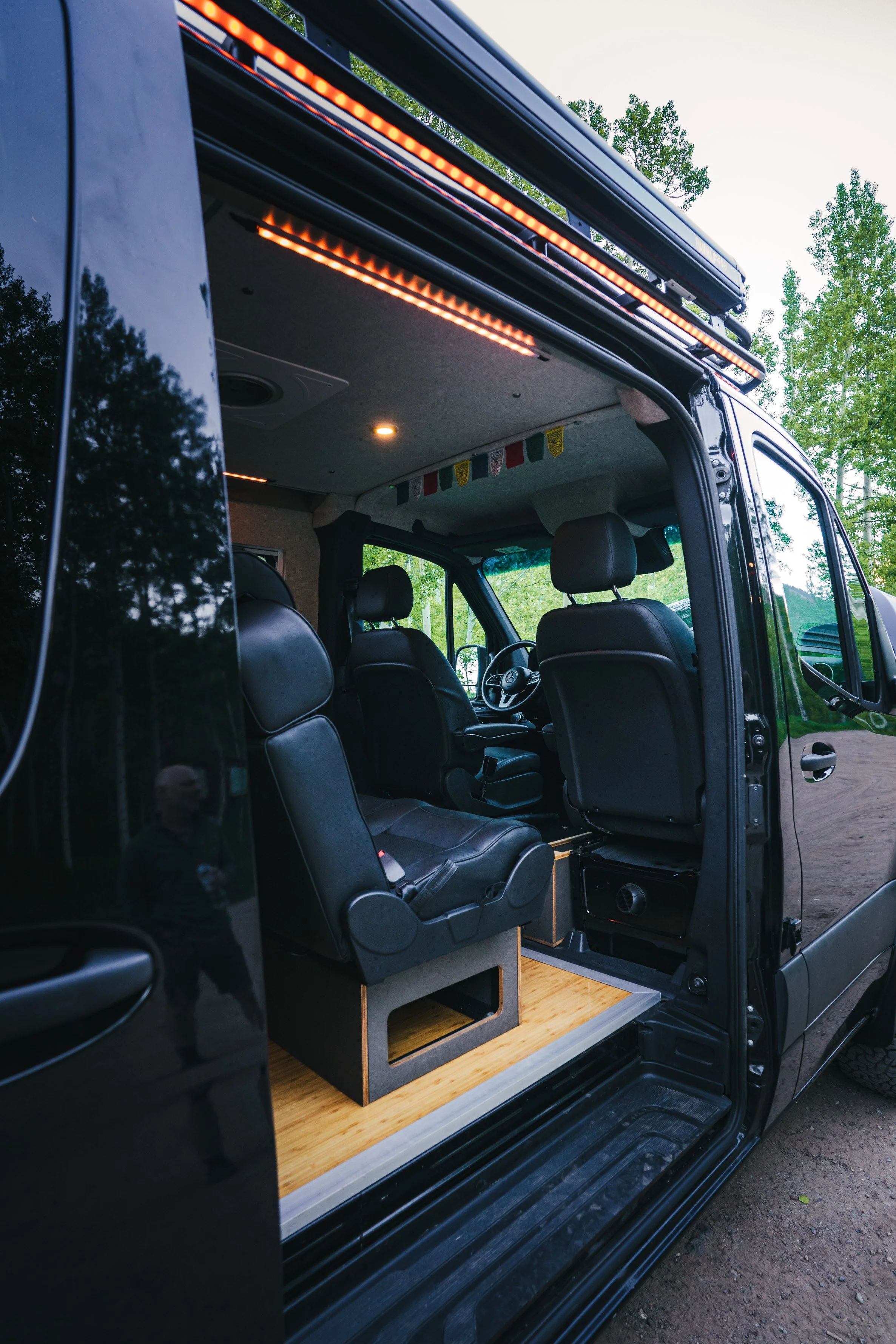 Interior of a black campervan with black leather seats, wooden flooring, and interior lighting, parked outdoors among green trees.