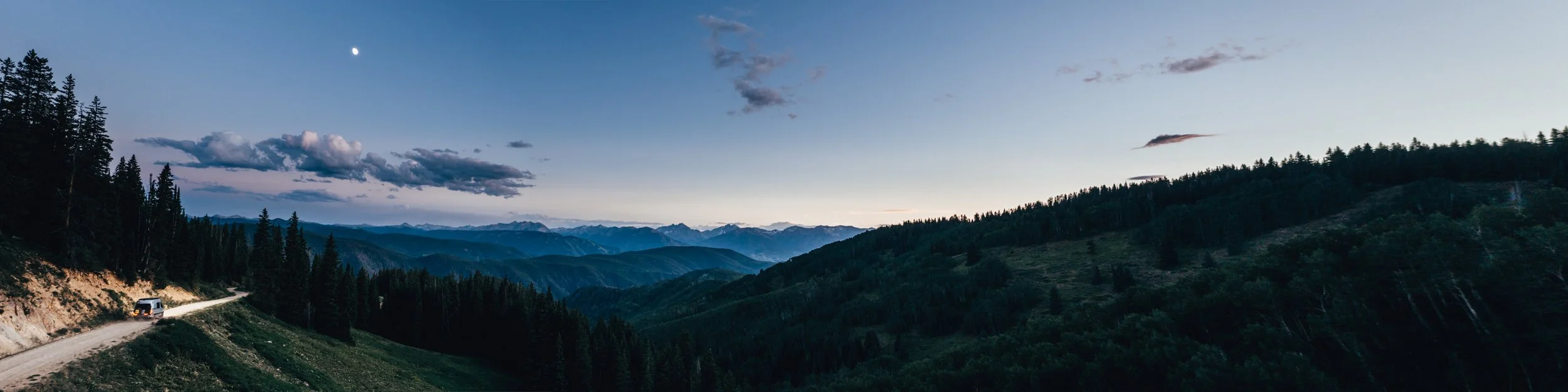 Scenic mountain landscape at dusk with a car driving on a gravel road, dense evergreen forests, rolling hills, and distant snow-capped mountains under a partly cloudy sky with a visible moon.