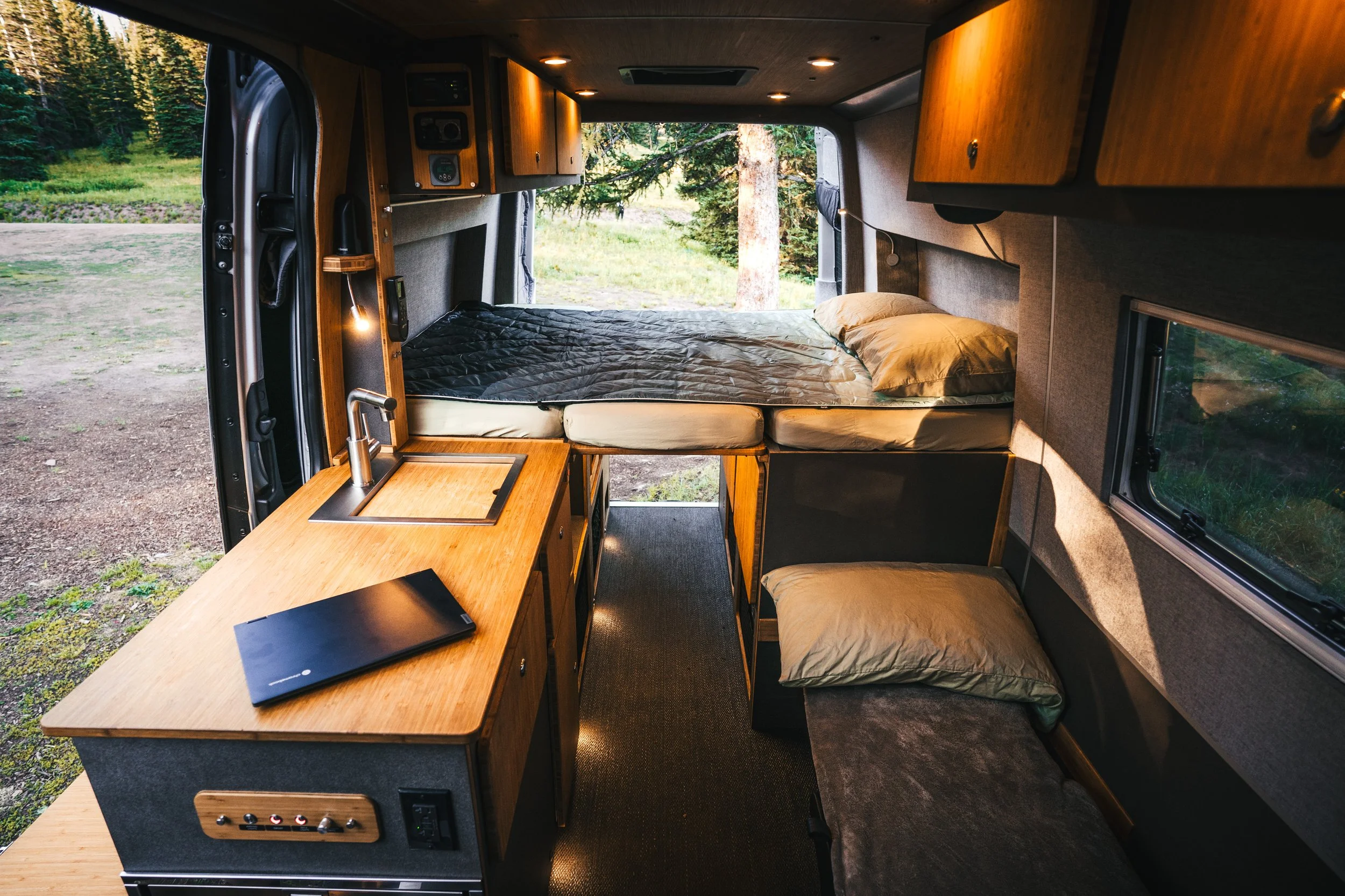 Interior of a camper van with a bed, small kitchenette with sink, and window overlooking a forested area.