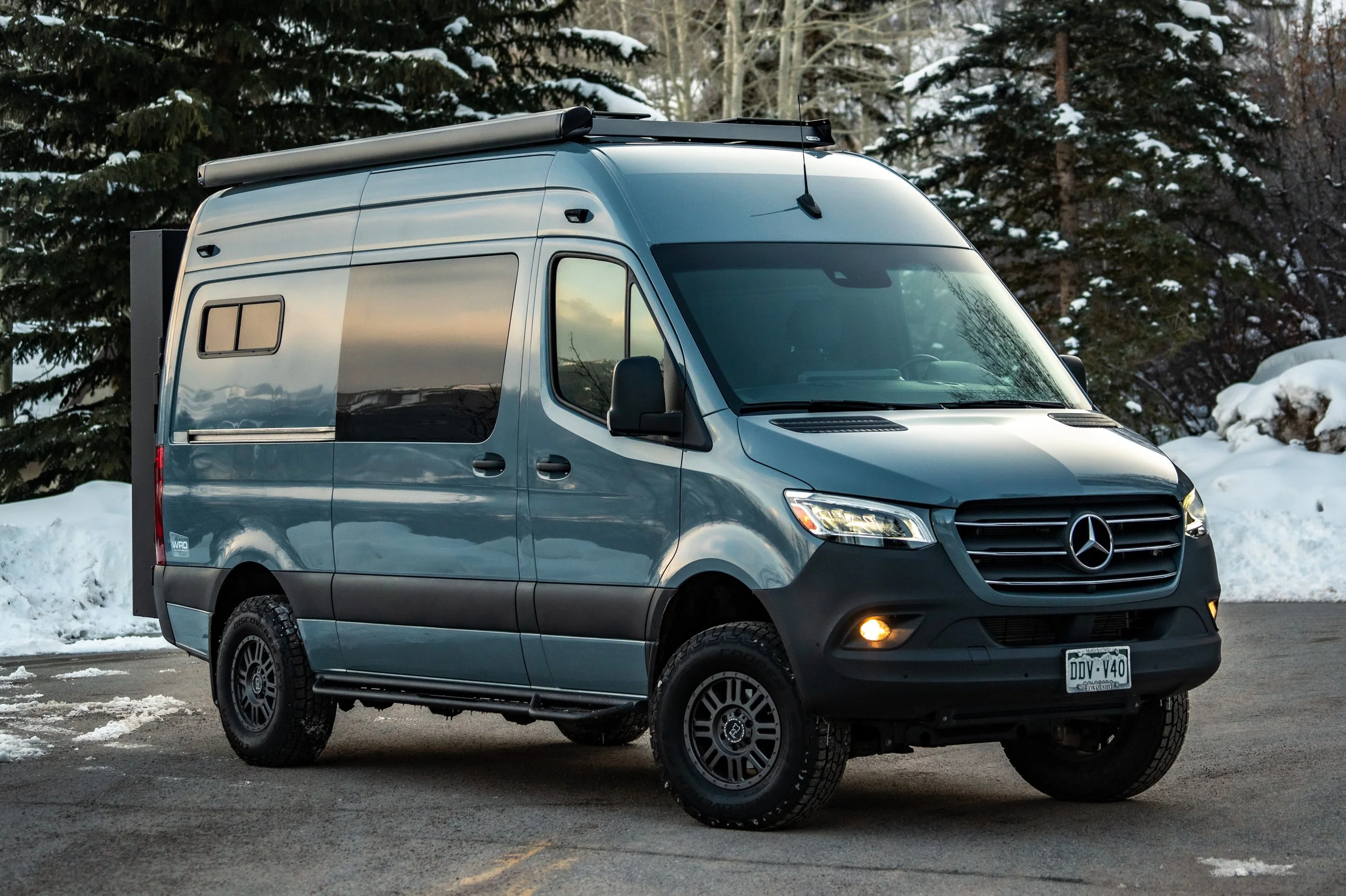 A blue Mercedes-Benz Sprinter camper van parked on a snowy road with snow-covered trees in the background.