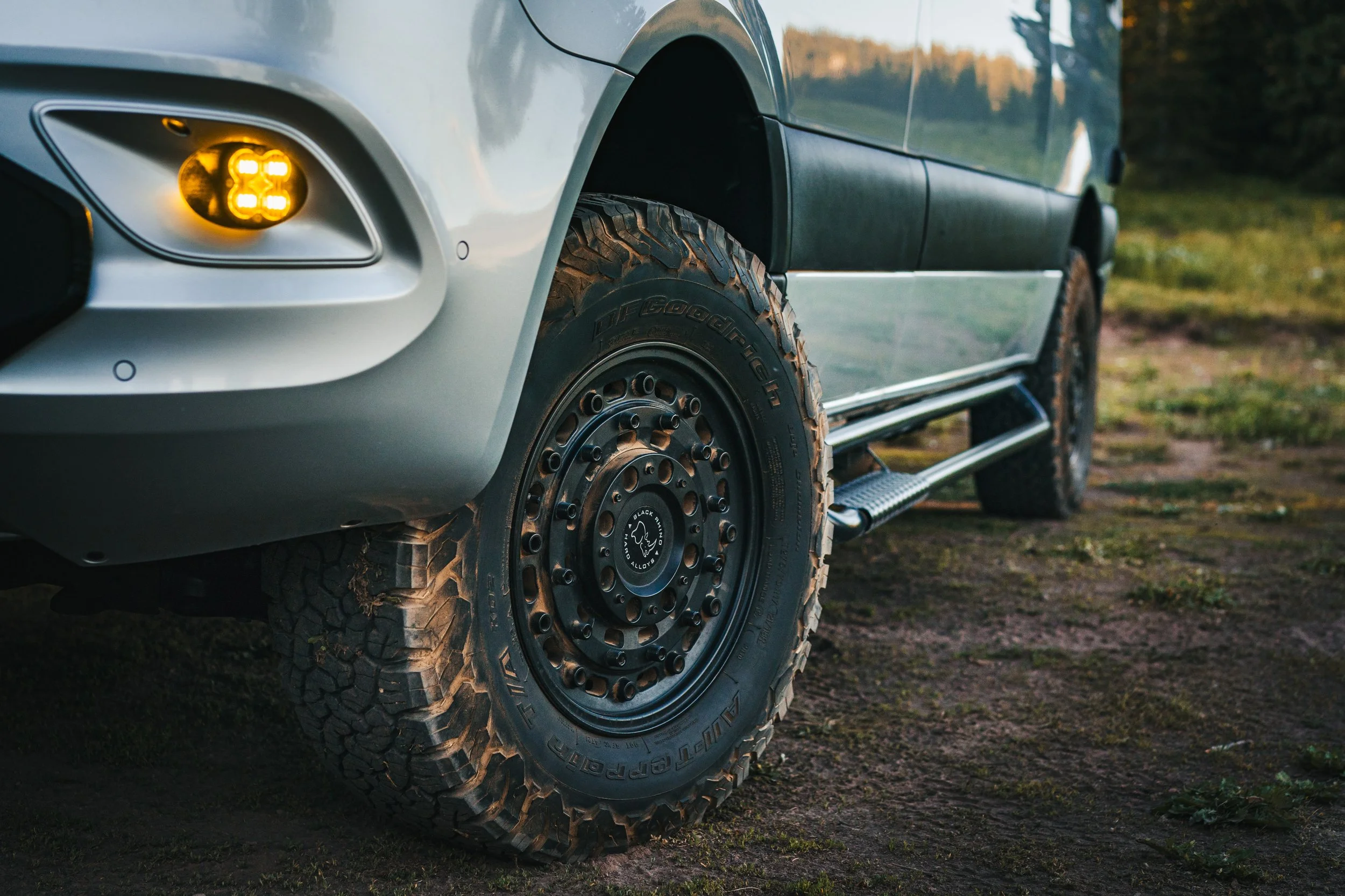 Close-up of a silver off-road vehicle with large, rugged tires, parked on a dirt field during sunset.