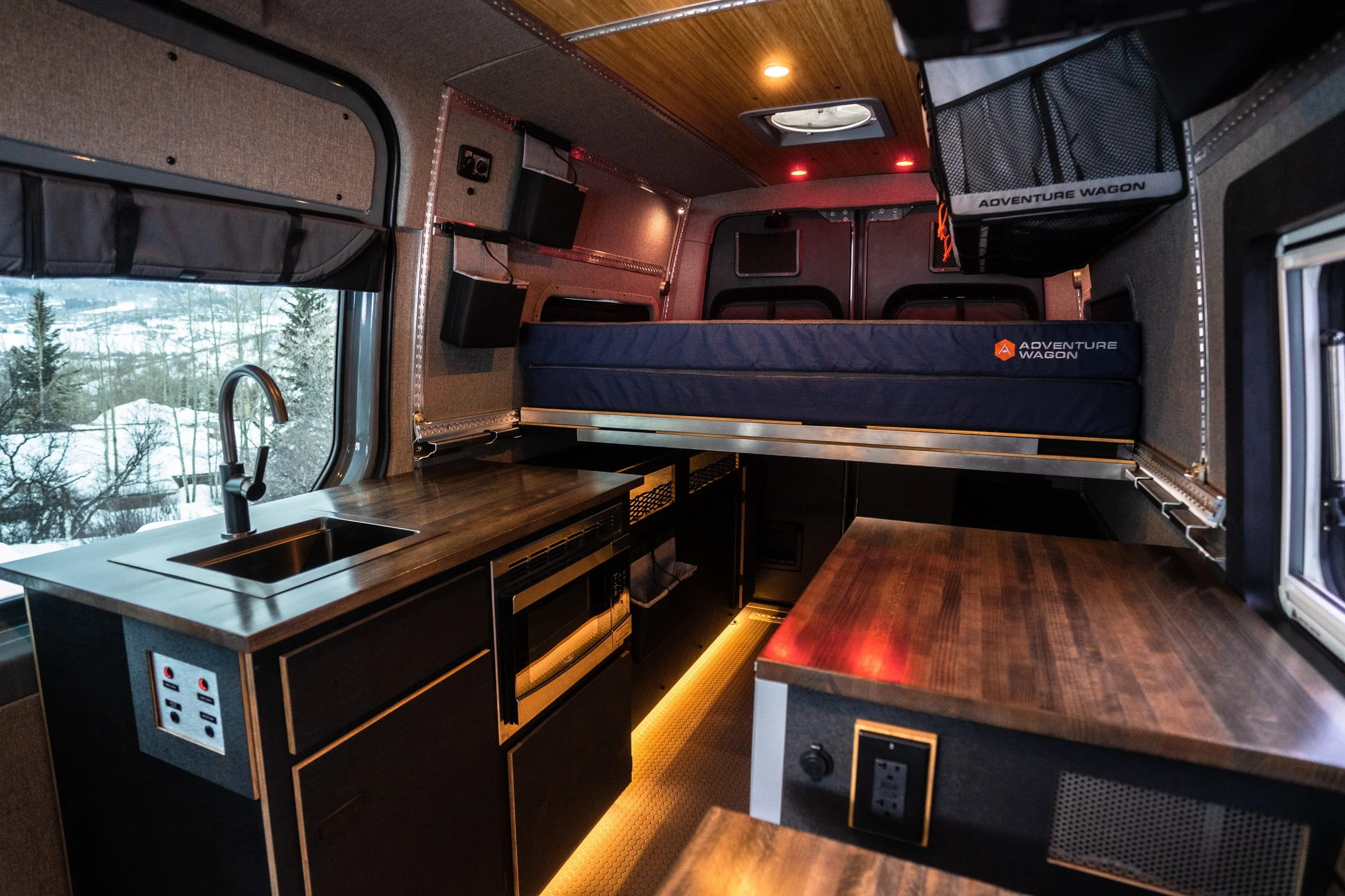 Interior of a compact camper van with a small kitchen area, a fold-out bed, and storage compartments.