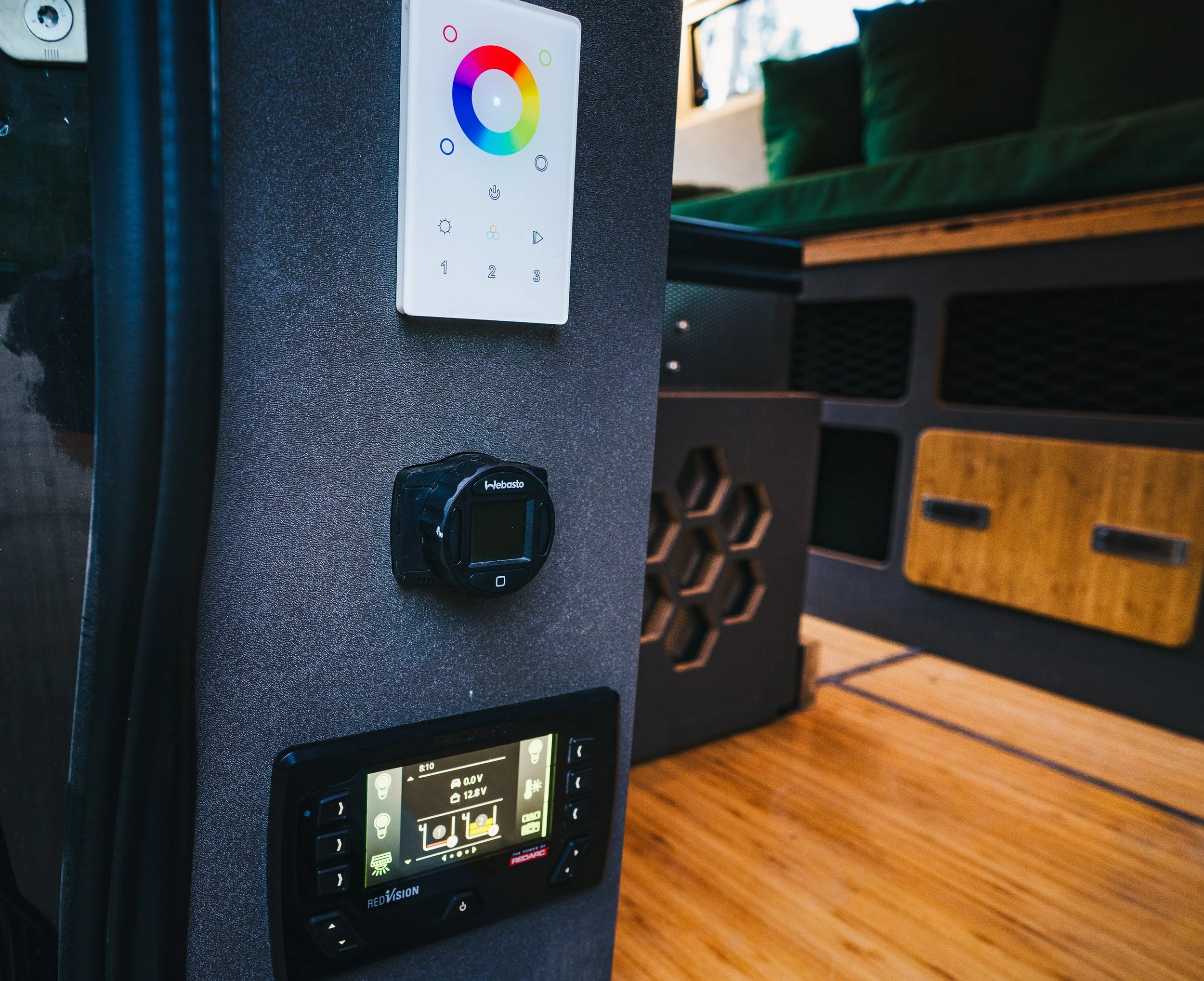 Close-up of a control panel with a color wheel remote, a black digital device, and an electronic display, set against a textured gray background. In the background, there is a wooden floor, a black and wooden cabinet, and a green couch.