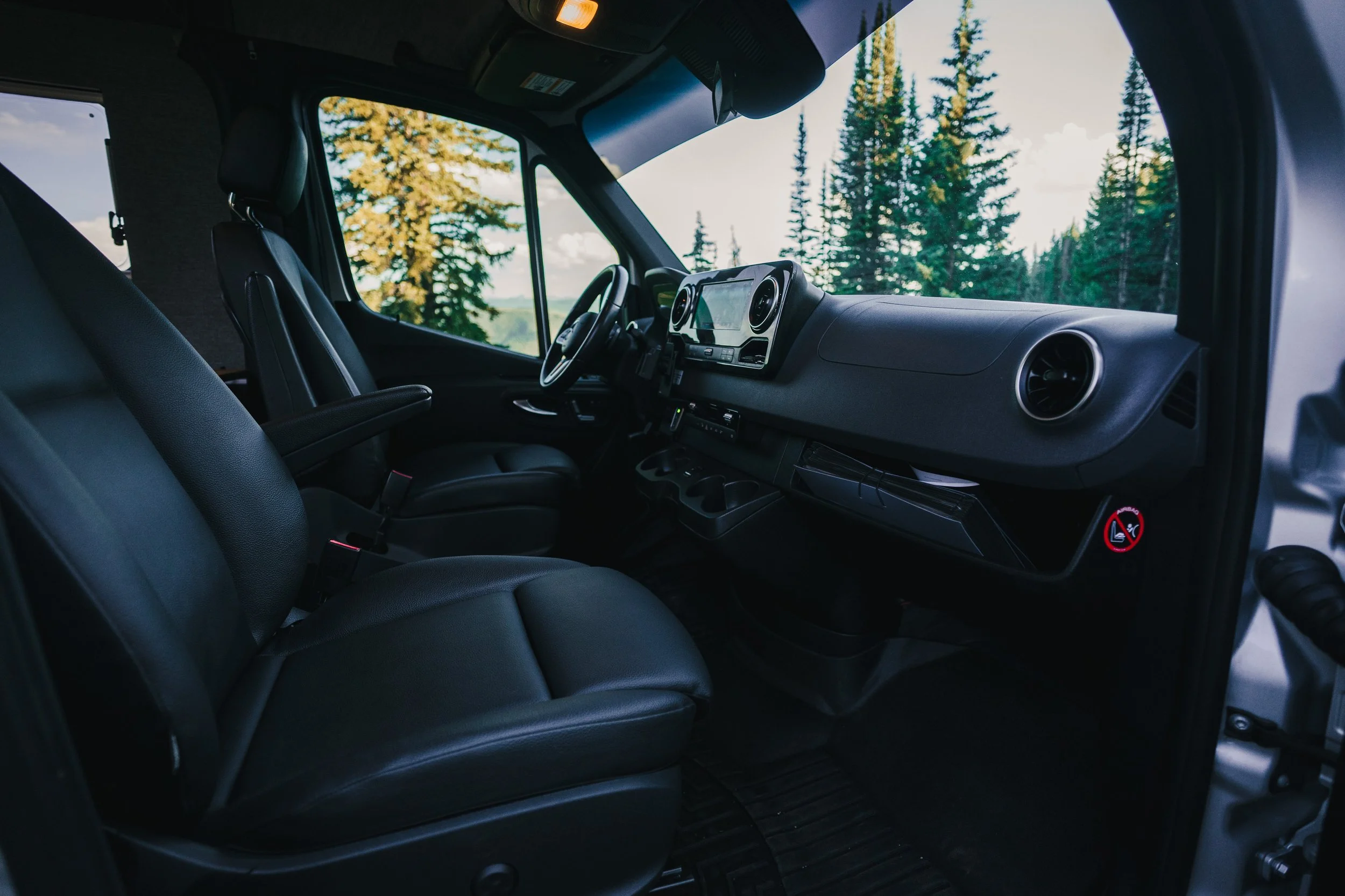 Interior view of a vehicle with black leather seats, dashboard, steering wheel, and a scenic view of trees outside through the window.