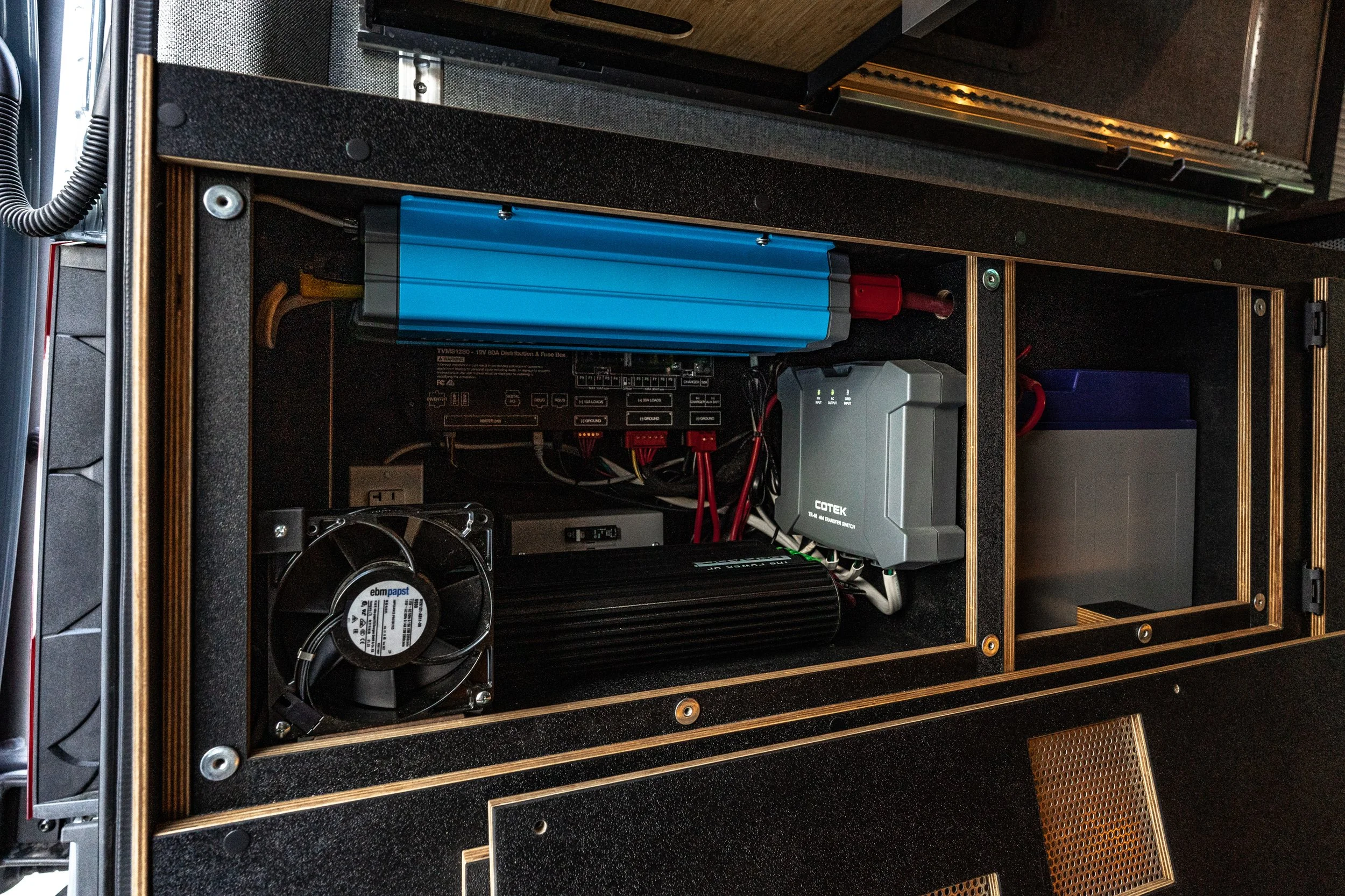 Inside a black wooden enclosure housing electronic components including a blue battery pack, cooling fan, power supply unit, and various cables and circuit boards.