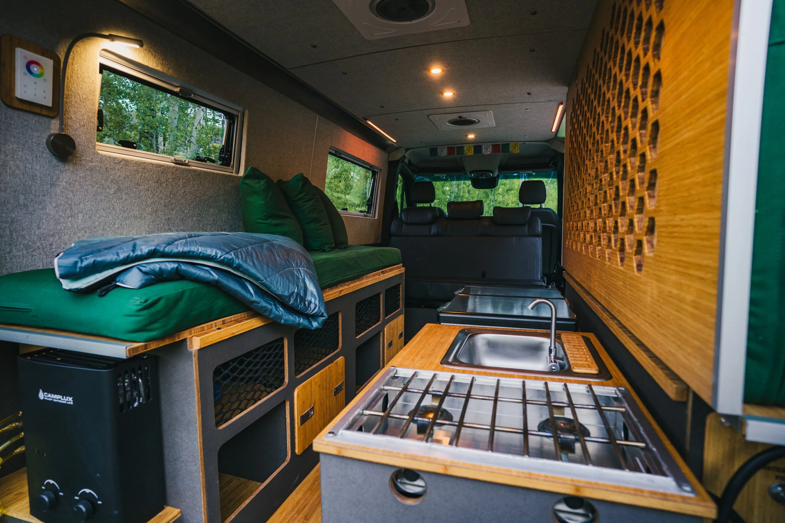 Interior of a camper van with a small kitchen, bed with green bedding, and windows with views of trees.