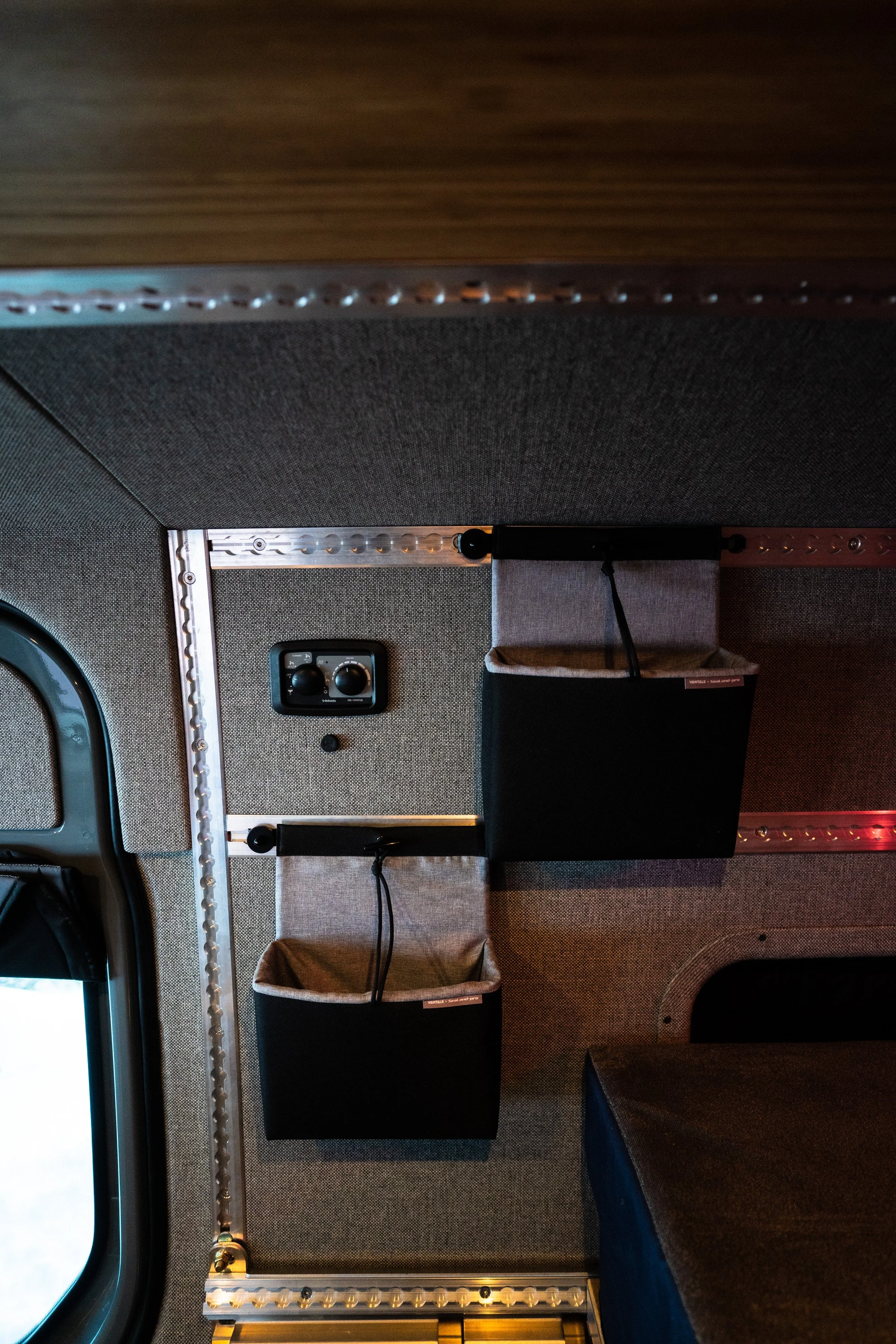 Interior of a vehicle with two storage bins, a black control panel, and exposed metal framing on the gray wall.