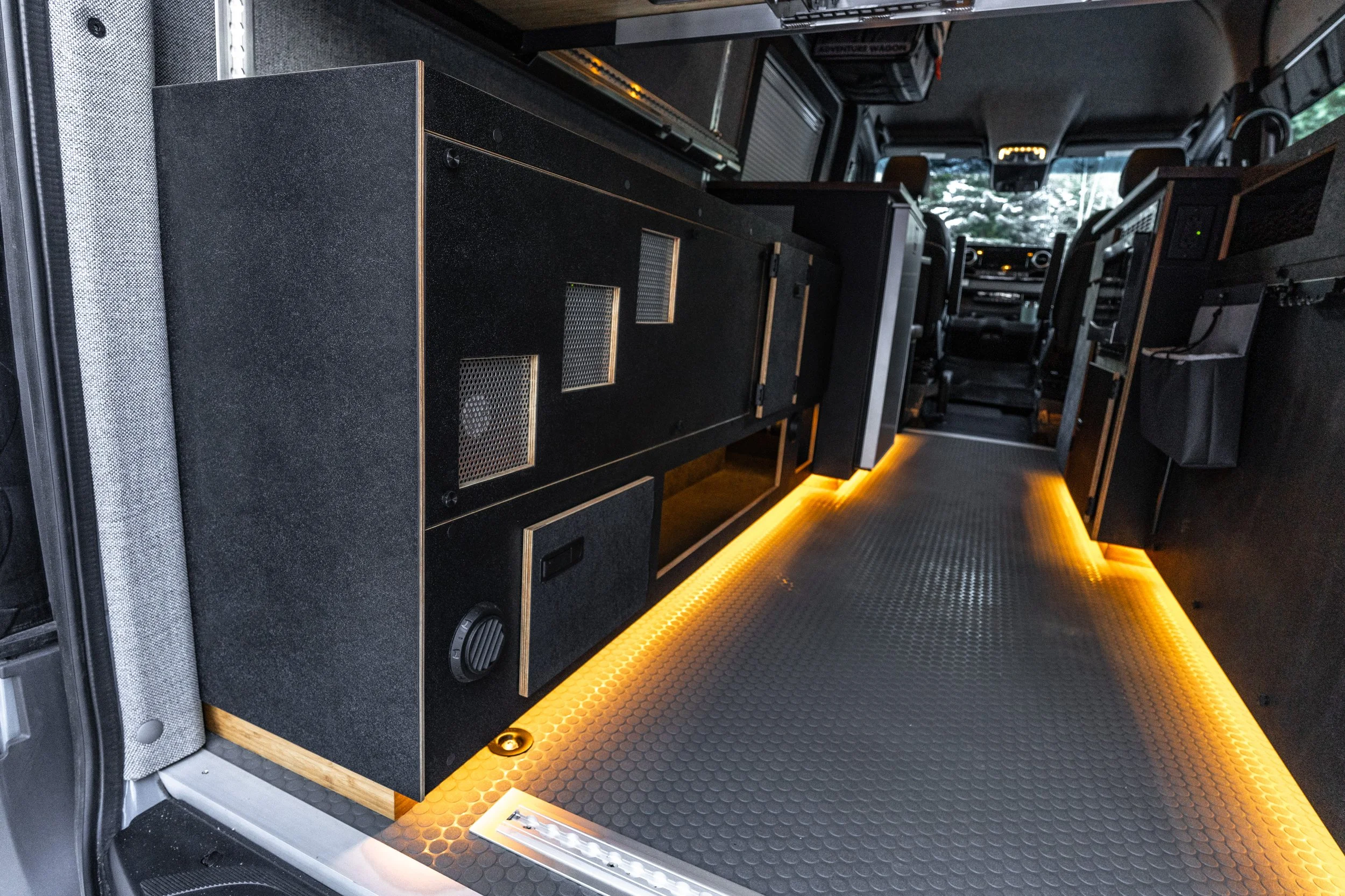 Interior of a van with dark cabinetry, illuminated yellow LED lighting on the floor, and the driver's cabin visible at the front.