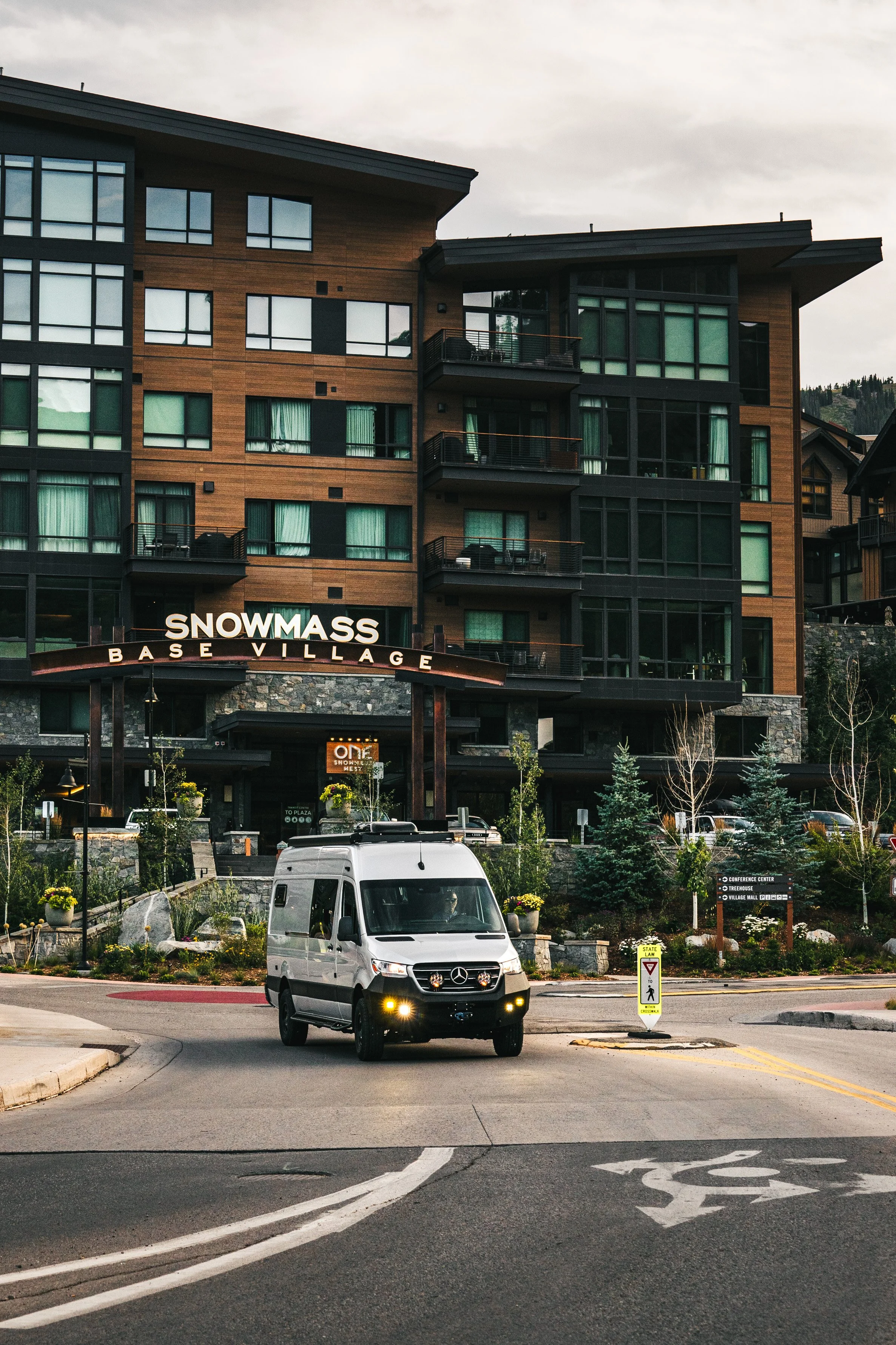 A street view of a modern multi-story hotel named Snowmass Base Village with a white Mercedes-Benz Sprinter van in the foreground, trees, and signs at the pedestrian crossing.