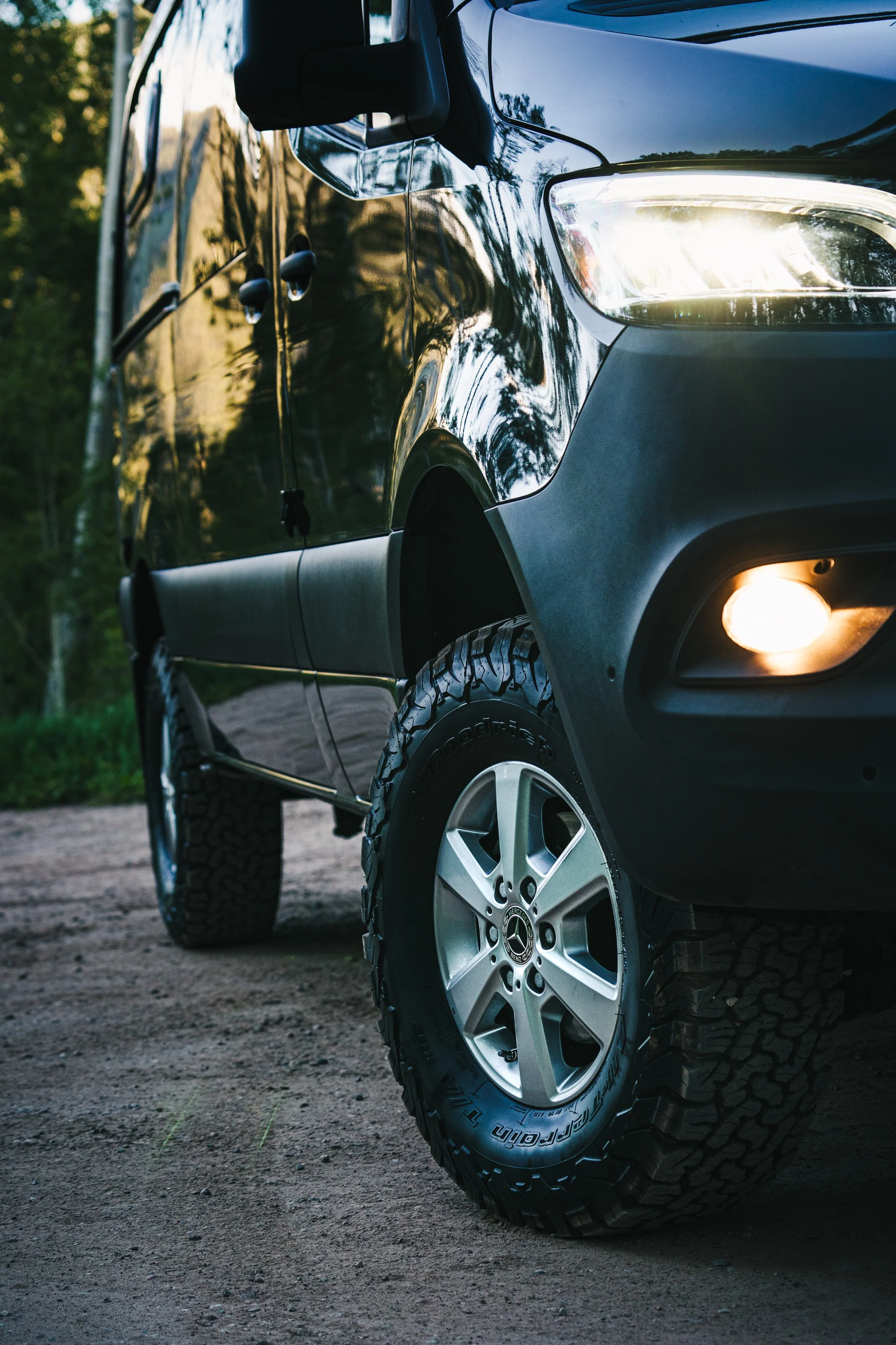 Close-up of a black SUV truck showing its front tire, headlight, and part of the hood, parked on a dirt road surrounded by trees.