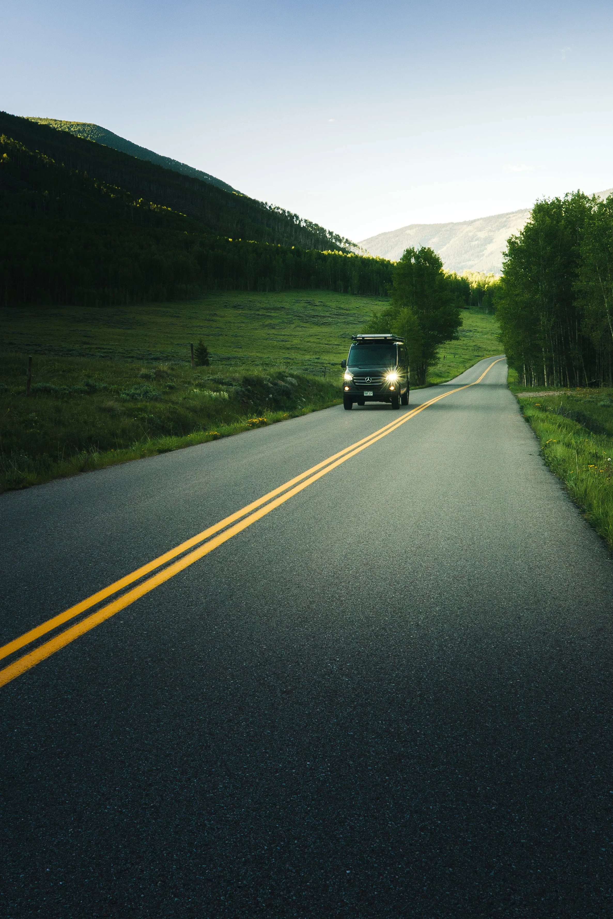 A black van driving on a two-lane rural road with double yellow lines, surrounded by green pastures and mountains in the background, under clear blue skies.